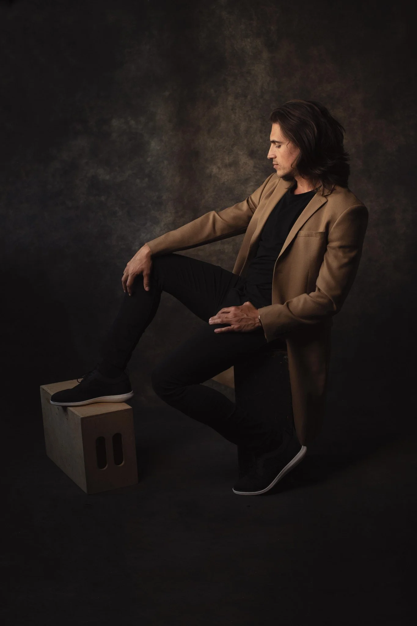 A man with long, dark hair wearing a tan blazer, black shirt, and pants, seated sideways on a chair with his right arm extended and his left arm resting on his lap, with one foot on a wooden block and the other foot on the ground, against a dark, tex