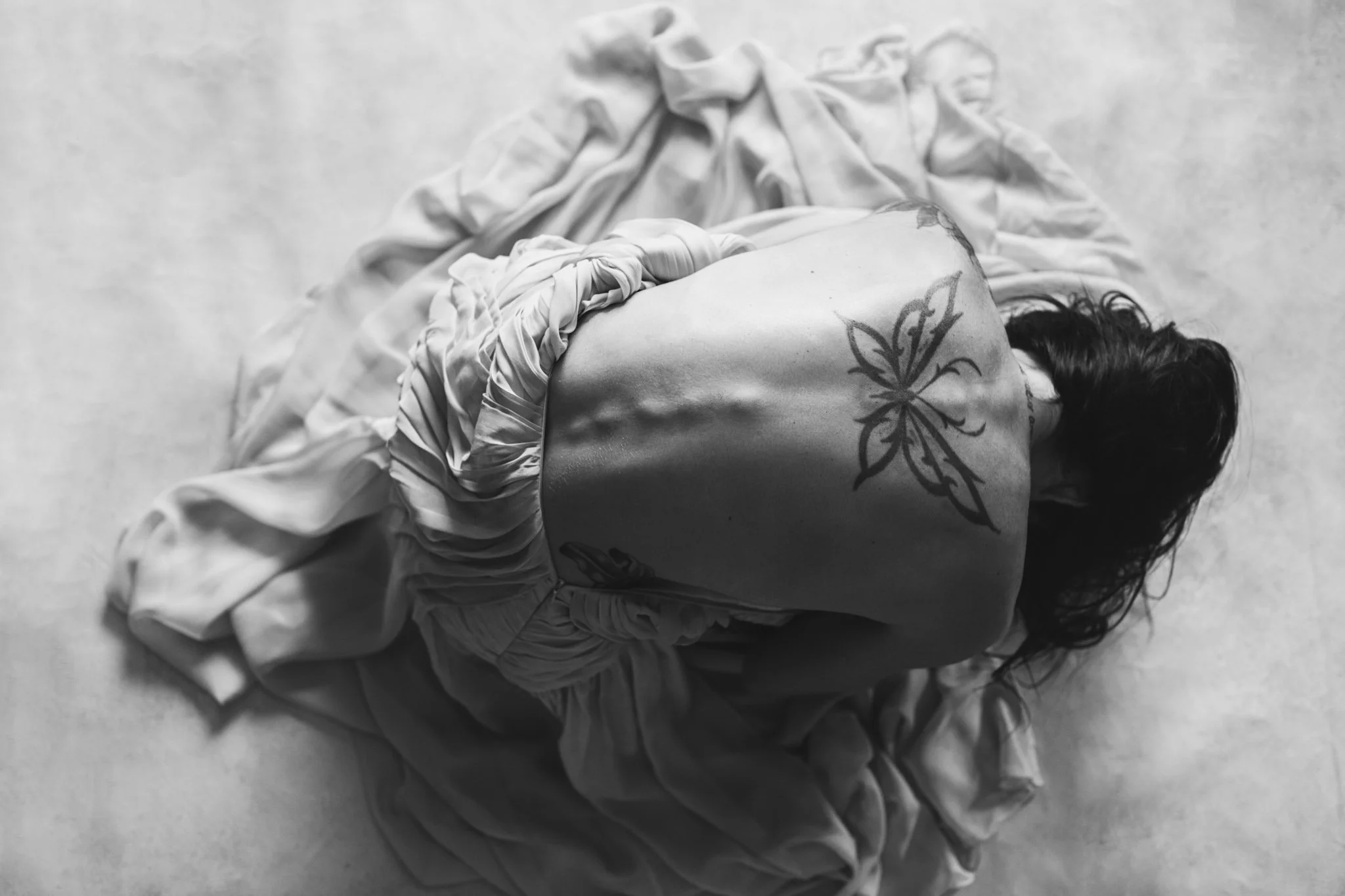 Top view of a woman with dark hair lying face down on a concrete floor, wrapped in light-colored fabric with one arm out, showing a butterfly tattoo on her shoulder and arm.