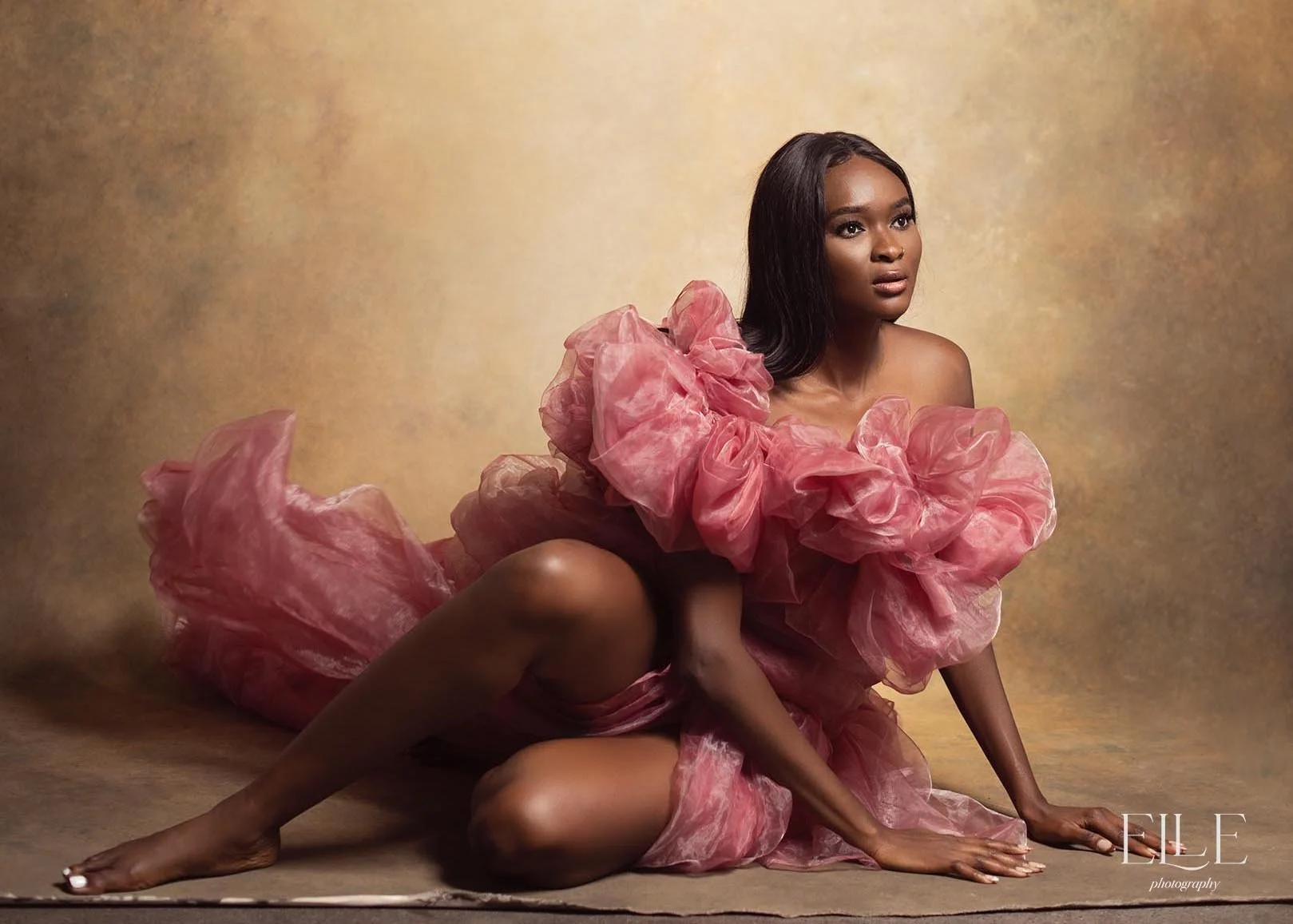 A woman sitting on the floor in a pink ruffled dress with a textured beige background.