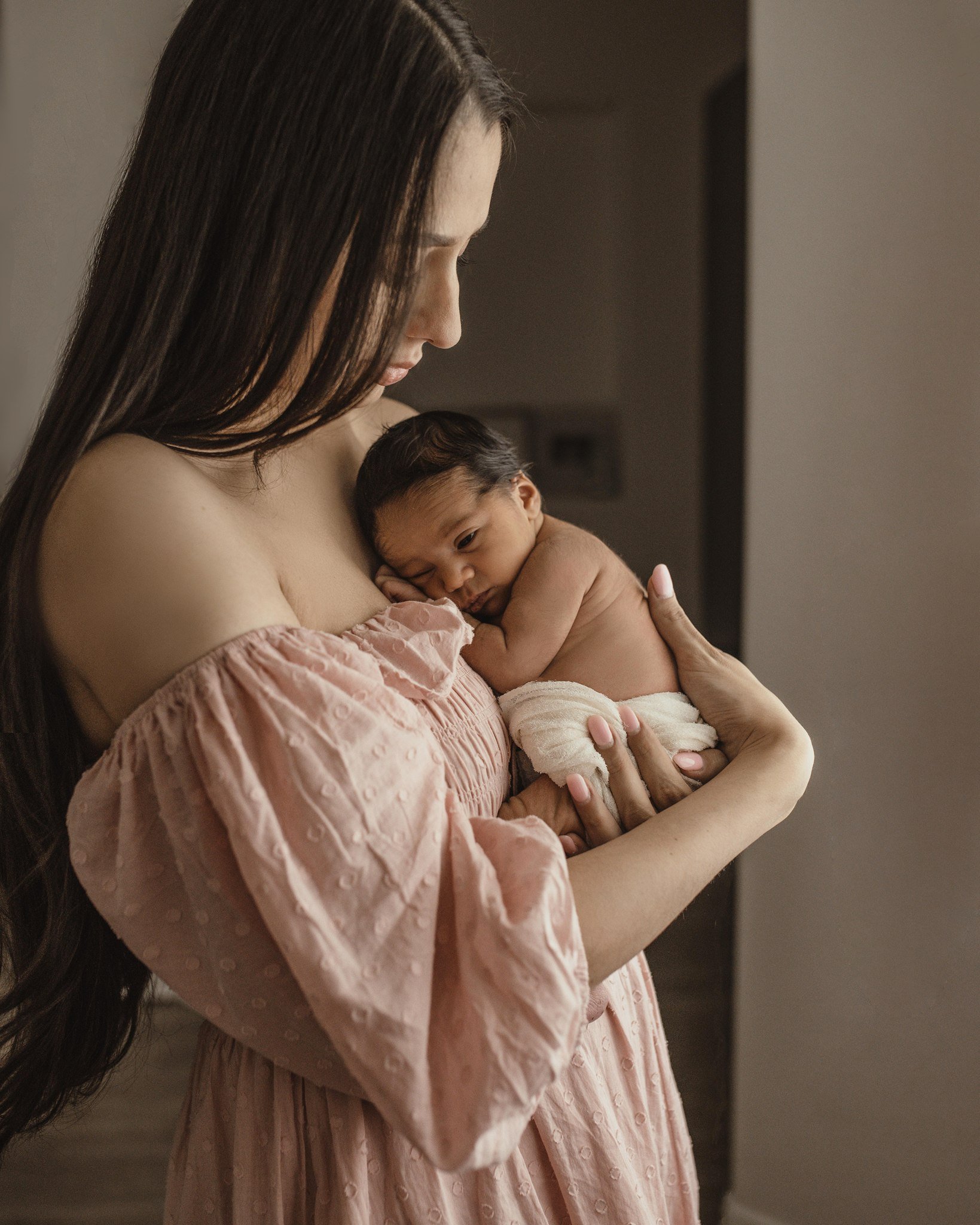 A woman with long dark hair holding a baby close to her chest. She is wearing a pink, textured dress with puffed sleeves. The baby has dark hair and is wrapped in a white blanket, resting peacefully. The background is neutral and slightly blurred.
