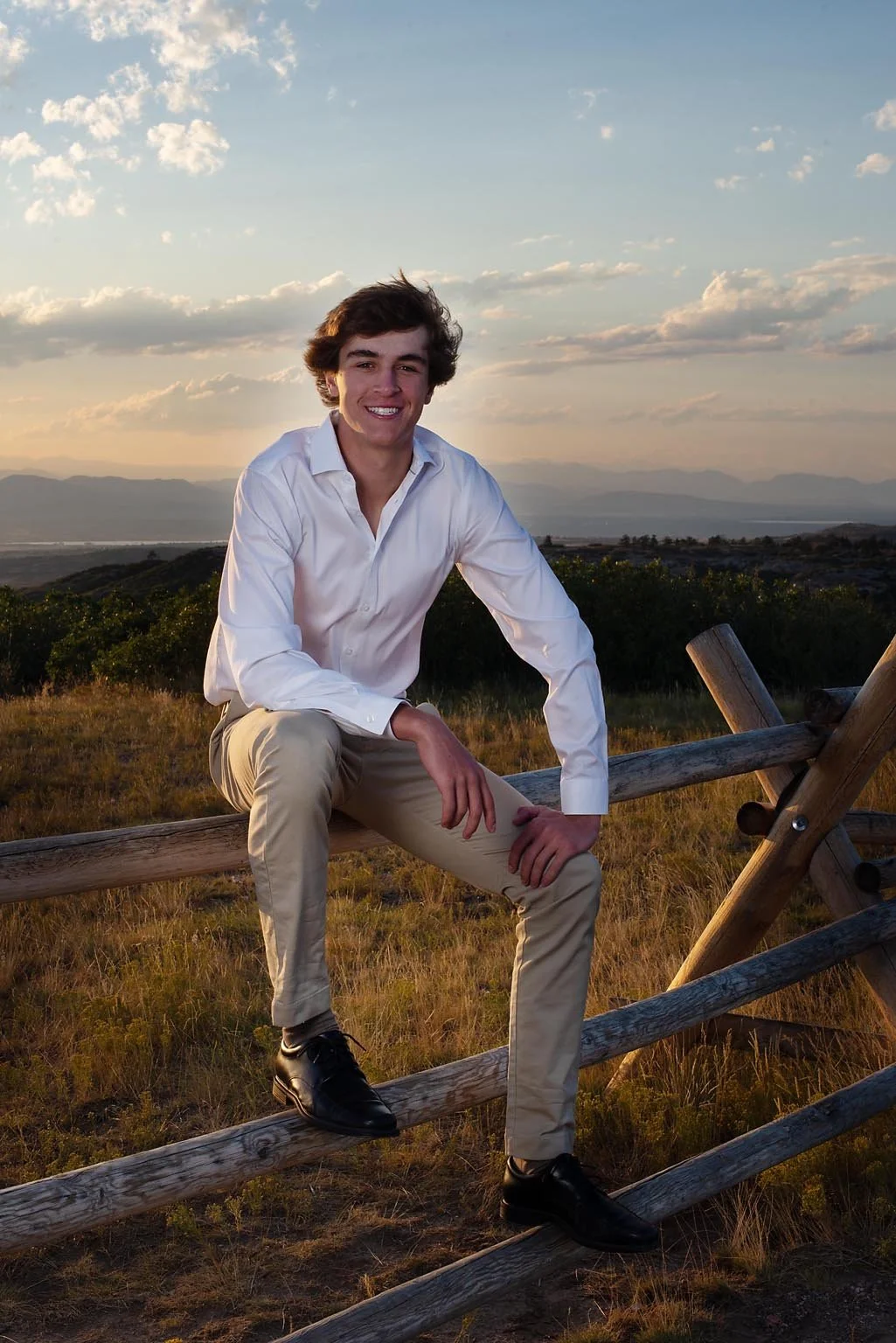 A young man with dark hair and a white dress shirt sitting on a wooden fence in an open field during sunset, with mountains and a sunset sky in the background.