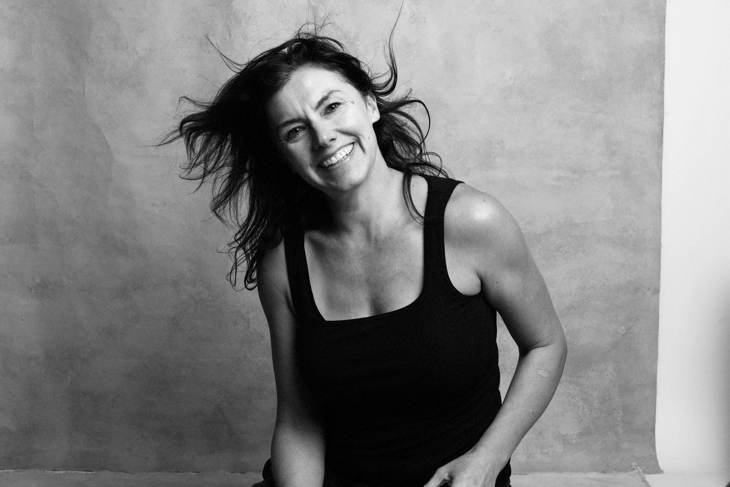 Black and white photo of a woman smiling, with hair flying, wearing a black sleeveless top, standing against a plain background.