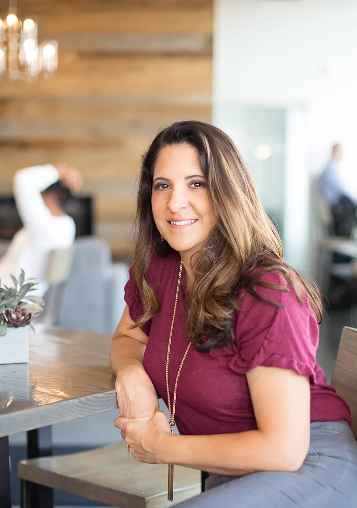 A woman with long brown hair, wearing a maroon top and a long necklace, sitting at a wooden table in a modern, well-lit cafe or restaurant, smiling at the camera with a blurred background of other people and wooden decor.