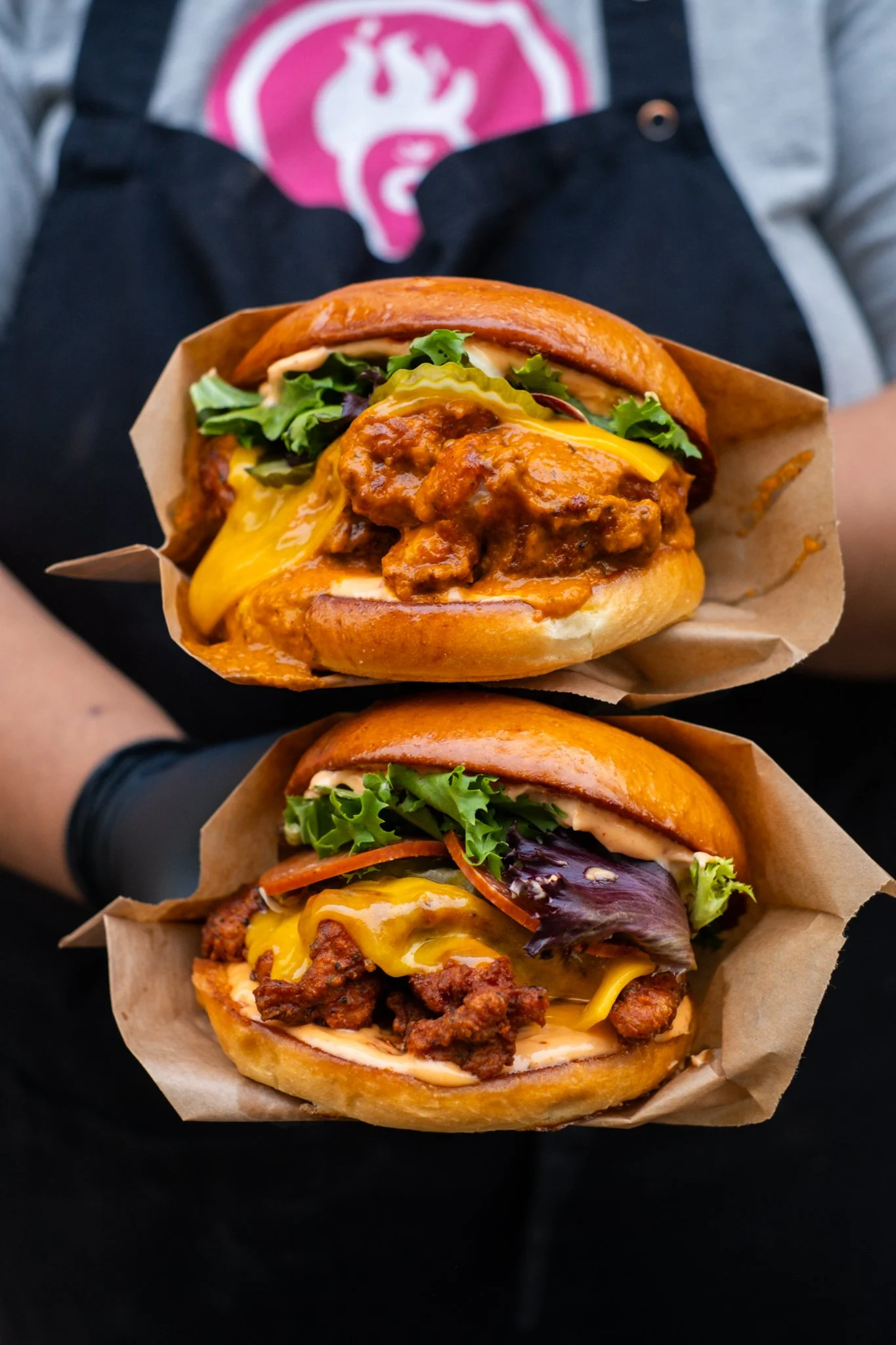Two burgers held in hands with brown paper wrappers, one topped with chili and cheese, the other with lettuce, tomato, and cheese, on toasted buns.