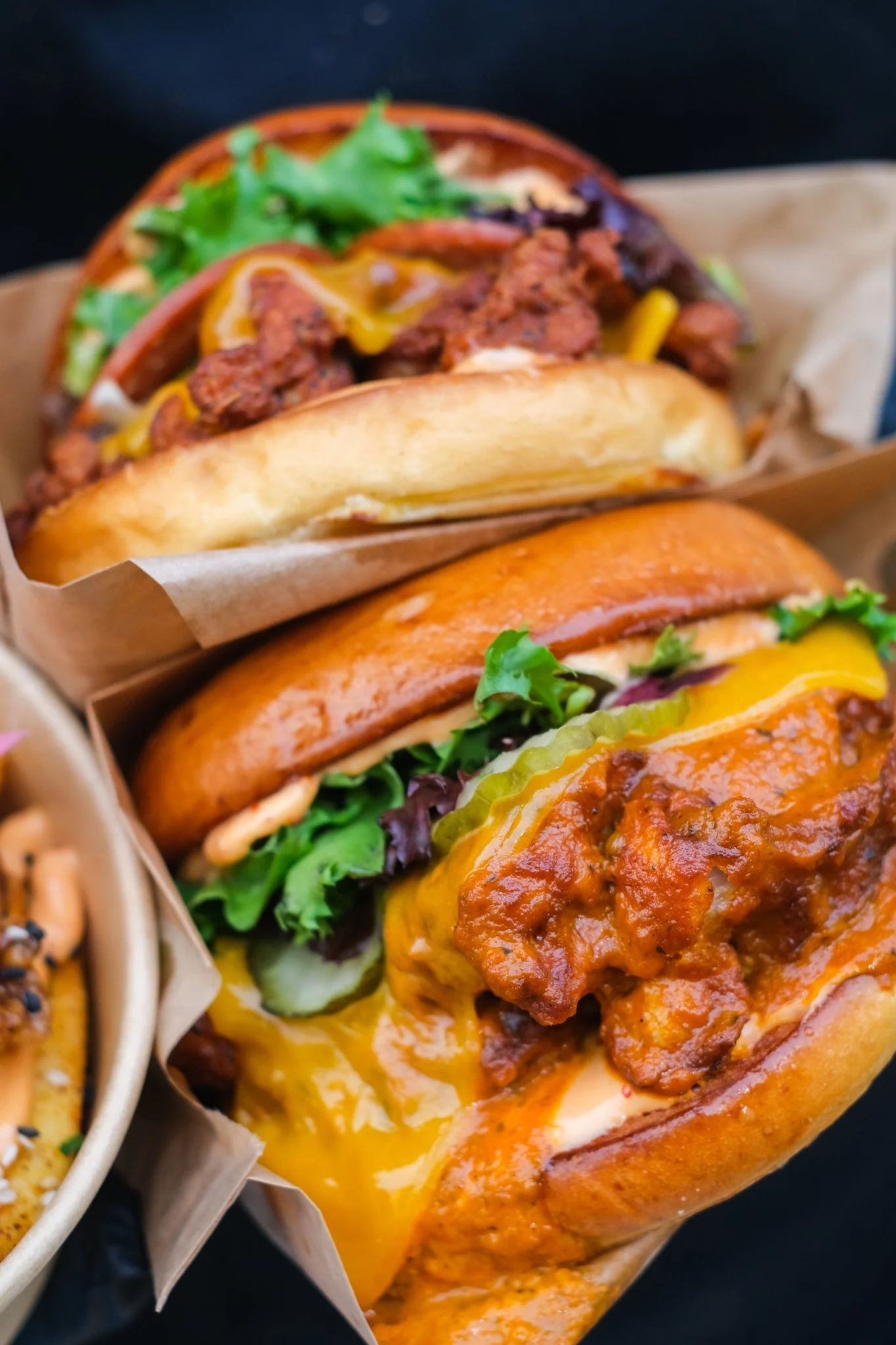 Two cheeseburgers with lettuce, pickles, and sauce in toasted buns, one with chili, served on brown paper-lined baskets.