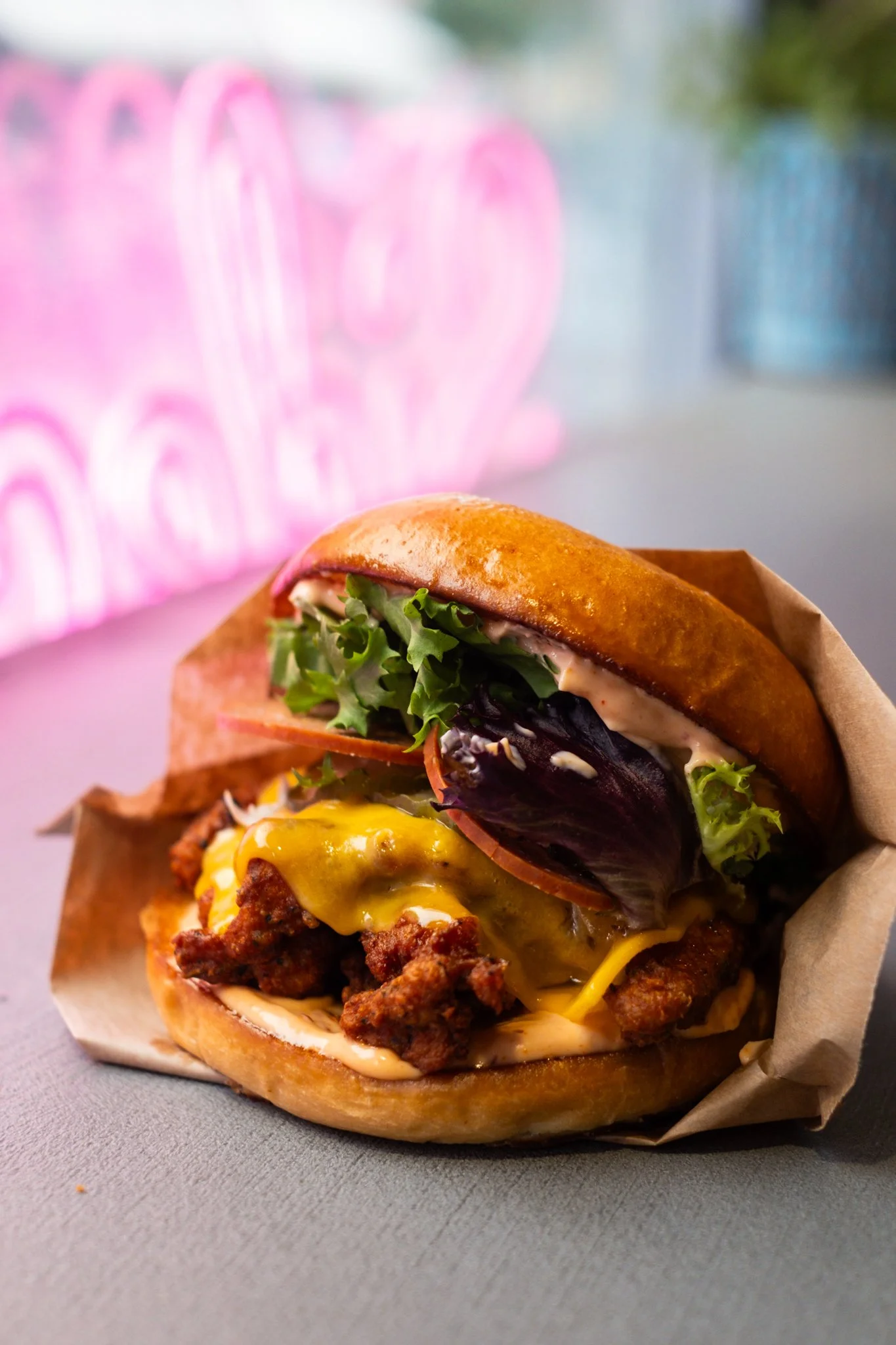 Close-up of a cheeseburger with fried chicken patty, melted cheese, lettuce, tomato, and sauce in a toasted bun, placed on a paper wrapper on a gray surface.