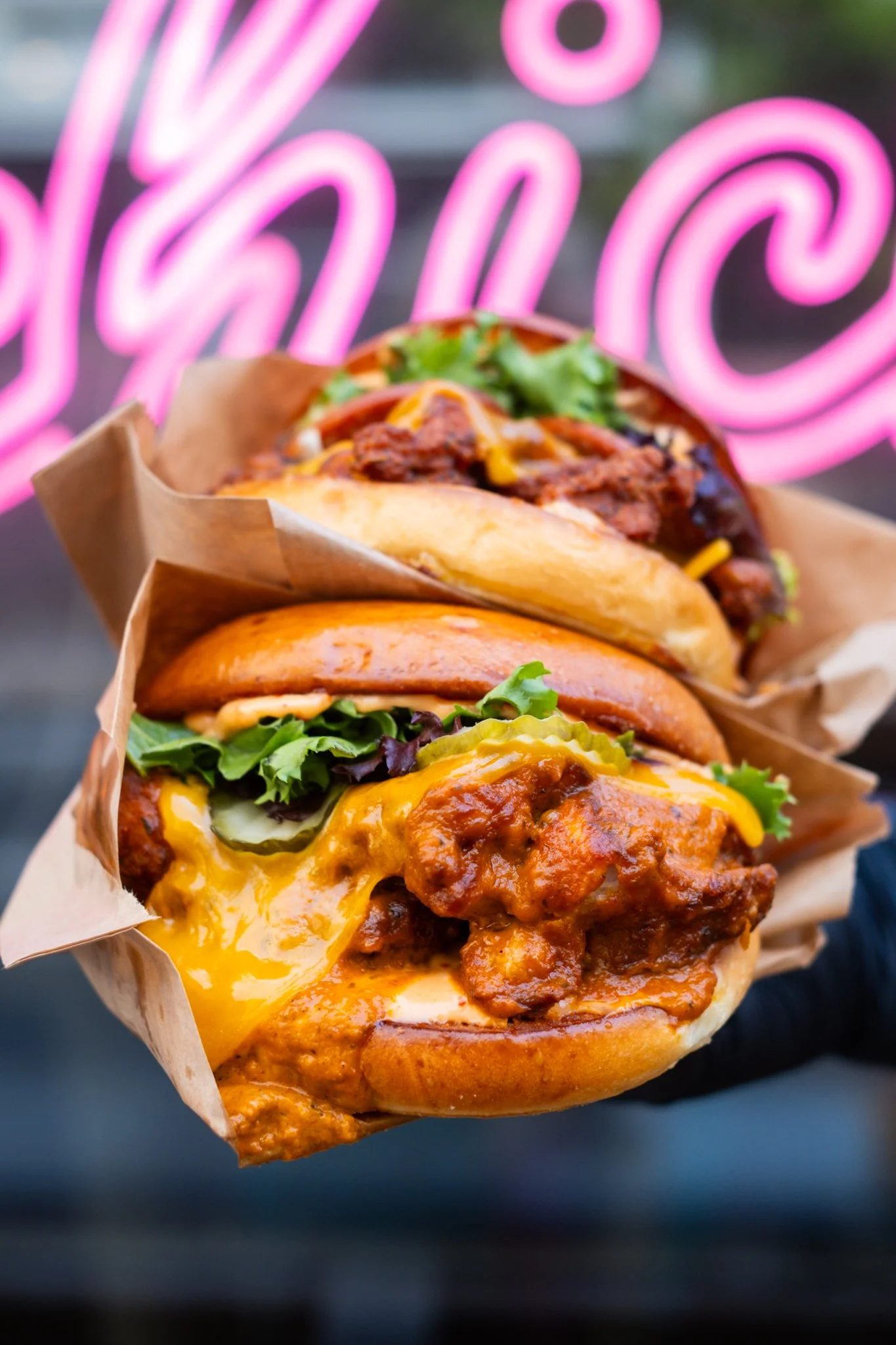 Two loaded burgers wrapped in brown paper, with cheese, lettuce, pickles, and sauces, held against a blurred background with pink neon lighting.