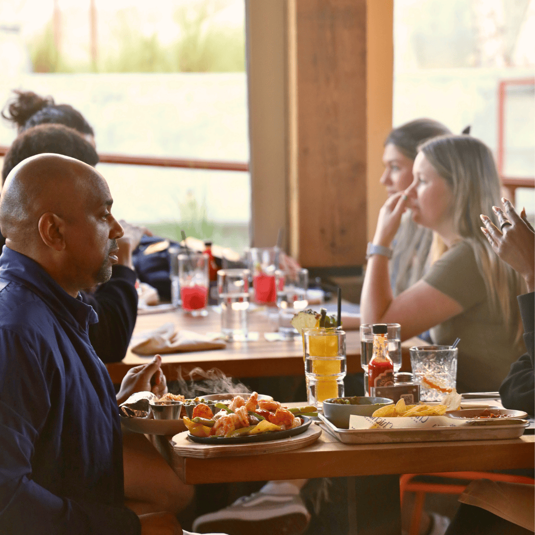 A man with a shaved head wearing a dark shirt is eating a meal with fried food at a restaurant. Several women sit at the table across from him, with drinks and condiments on the table.