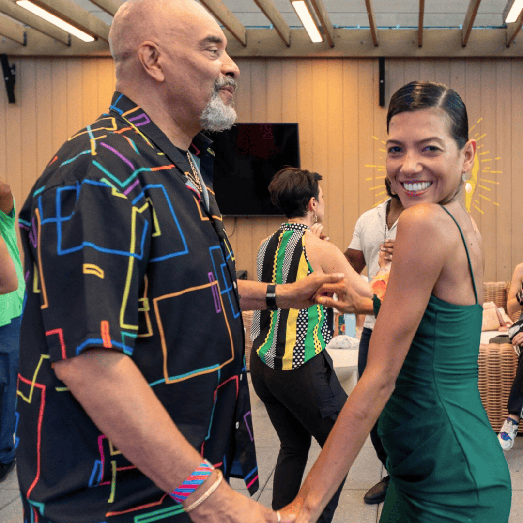 A man and woman dancing at a social gathering, holding hands, smiling, in a decorated indoor space with wood-paneled walls and other people in the background.