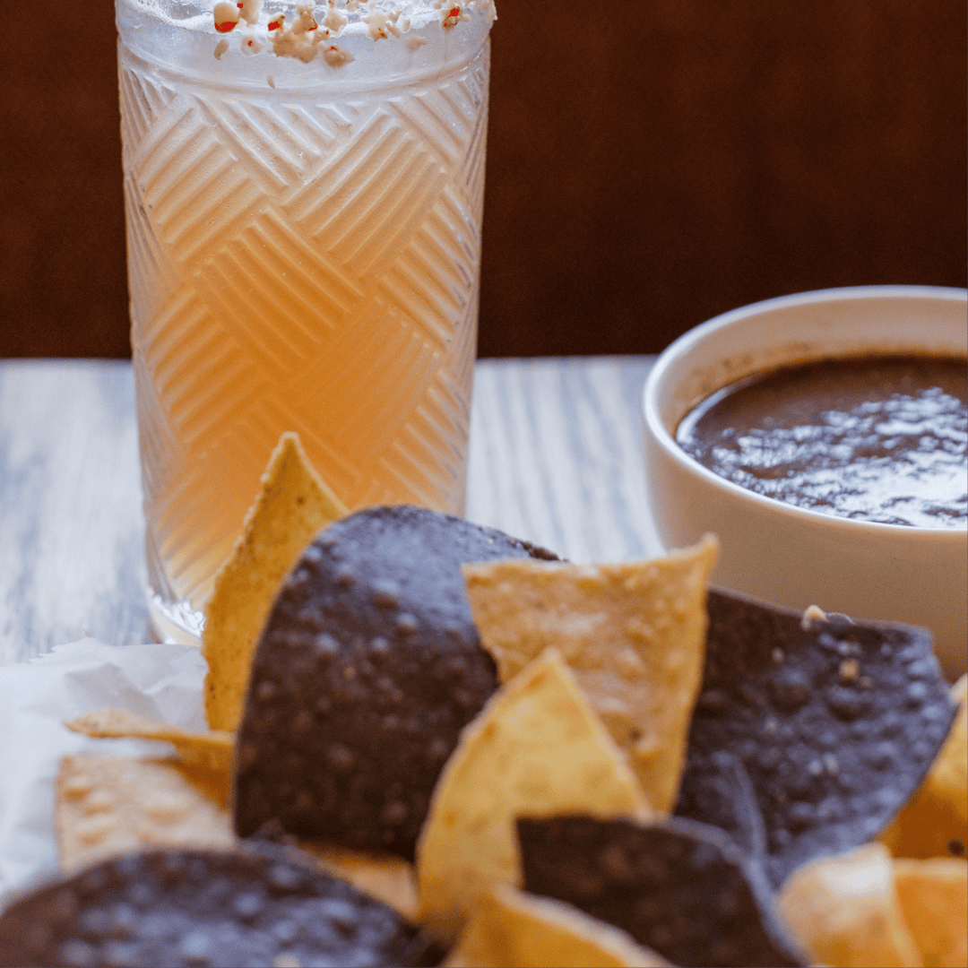 A glass of orange beverage topped with whipped cream and sprinkles, a bowl of chocolate sauce, and a pile of flavored tortilla chips in front of a wooden surface.