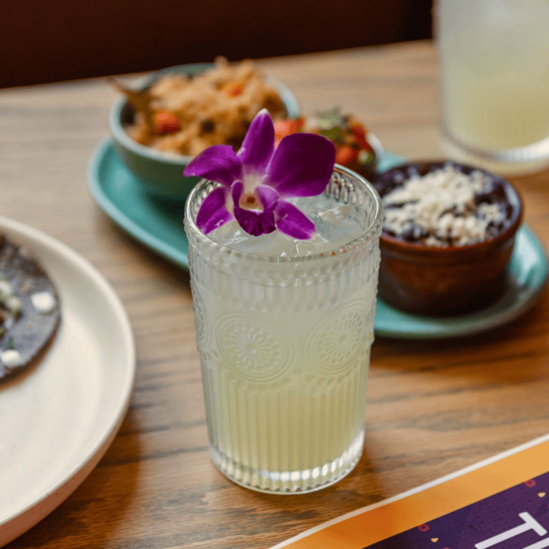 A tall glass of light-colored lemonade with an orchid flower on top, placed on a wooden table with various dishes in the background.