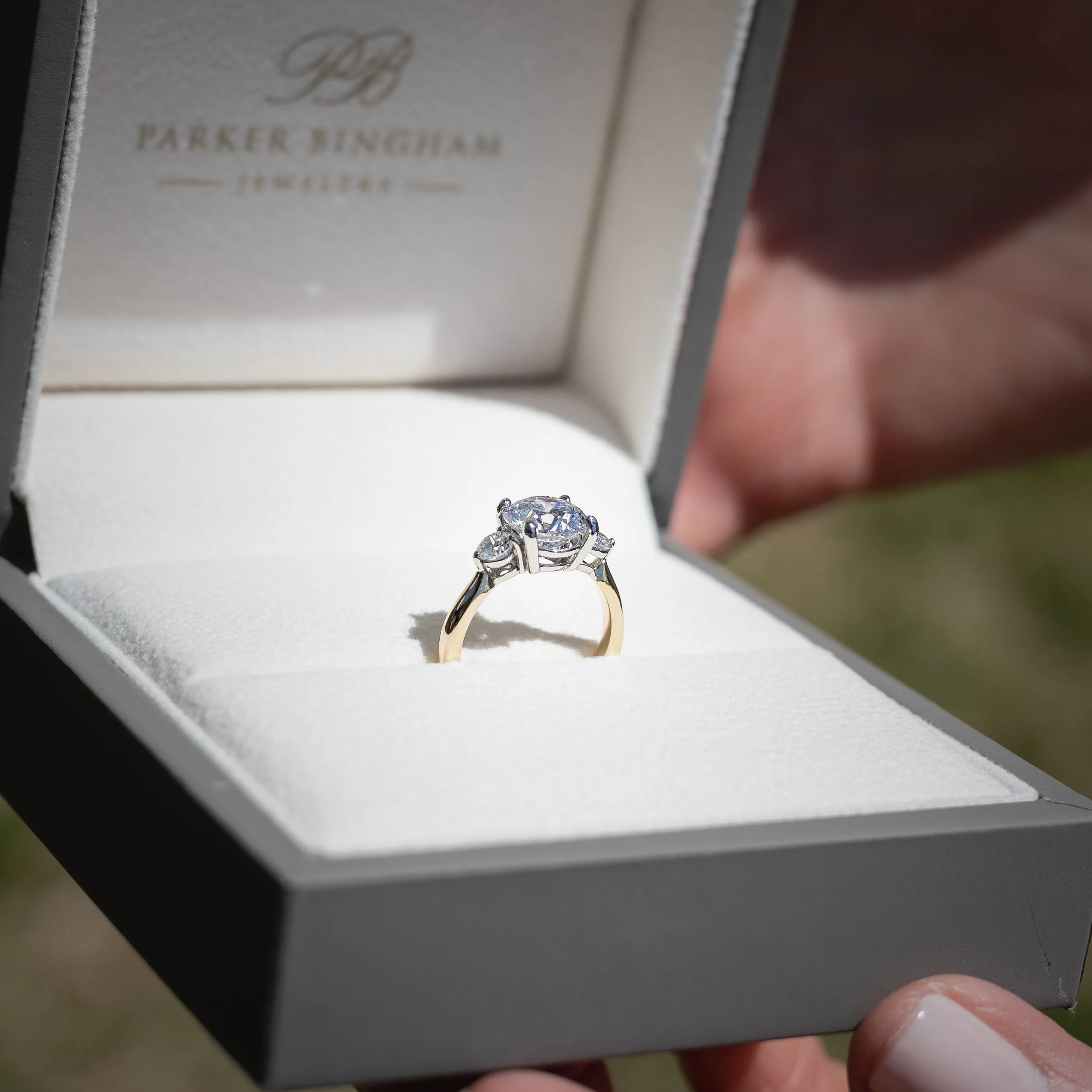 Close-up of a ring with a large central diamond with smaller diamonds around it, displayed in a white jewelry box with the brand name Parker Bingham Jewelry embossed inside.