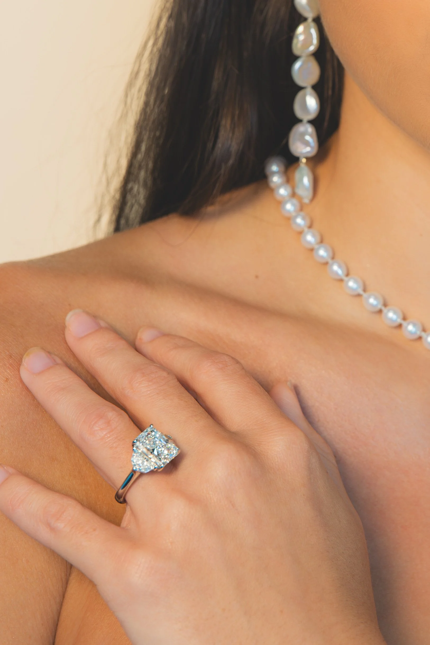 Close-up of a woman's hand with a large diamond engagement ring, placed on her shoulder. She is wearing a pearl necklace and diamond earrings.