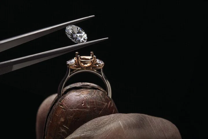 A diamond ring with a gold setting held by tweezers above a wooden object, with a loose diamond nearby.