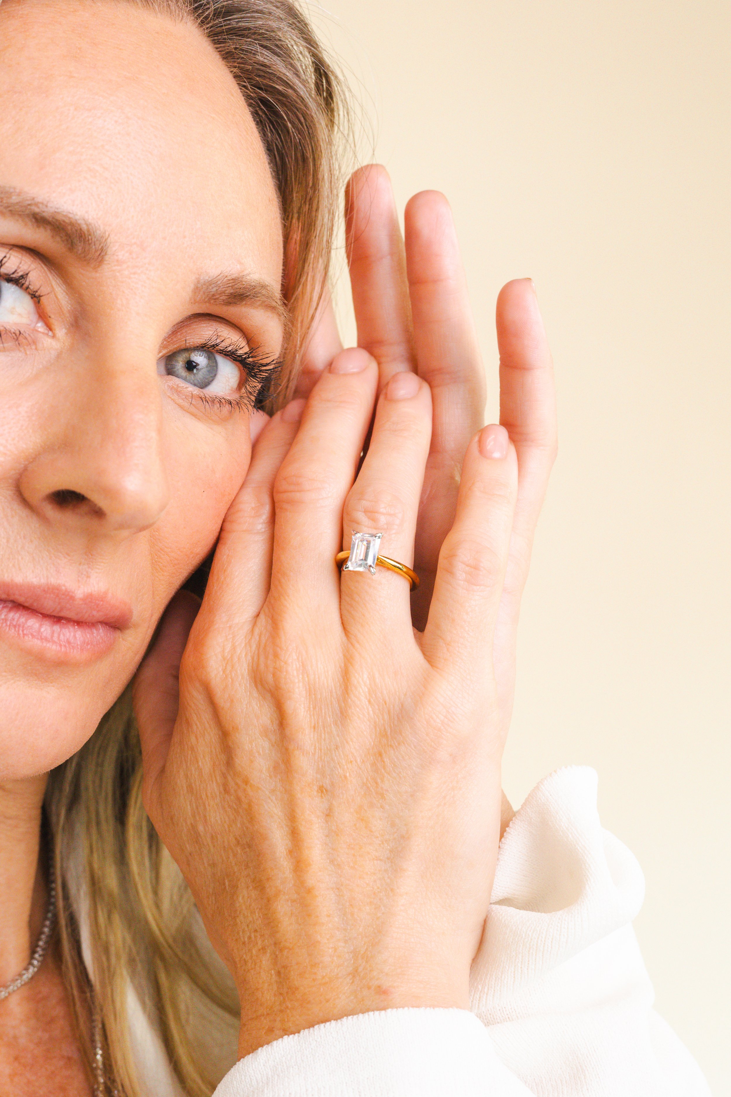 Close-up of a woman touching her face near her eye, wearing a white sweatshirt and a ring with a rectangular gemstone.