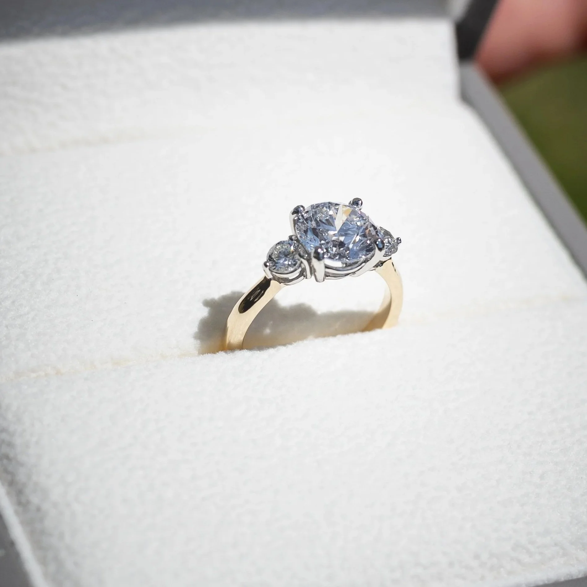 Close-up of a diamond engagement ring with a large central stone and smaller surrounding stones, set in a silver band, resting on a white surface.