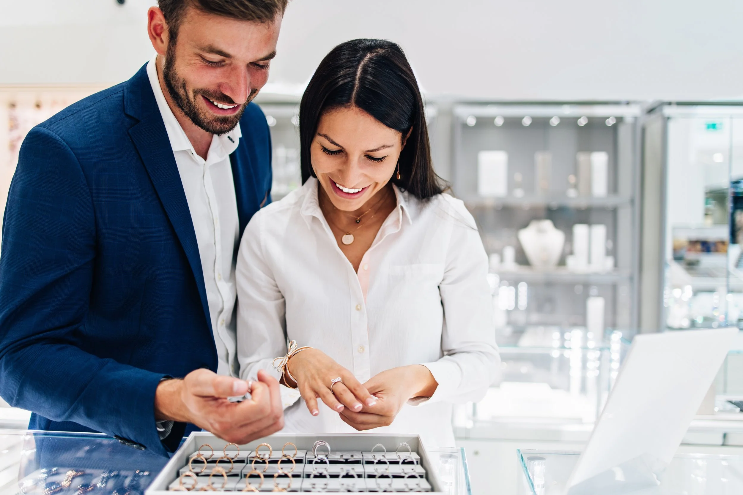 A man and woman shopping for jewelry, looking at rings and wearing smiles, in a jewelry store.