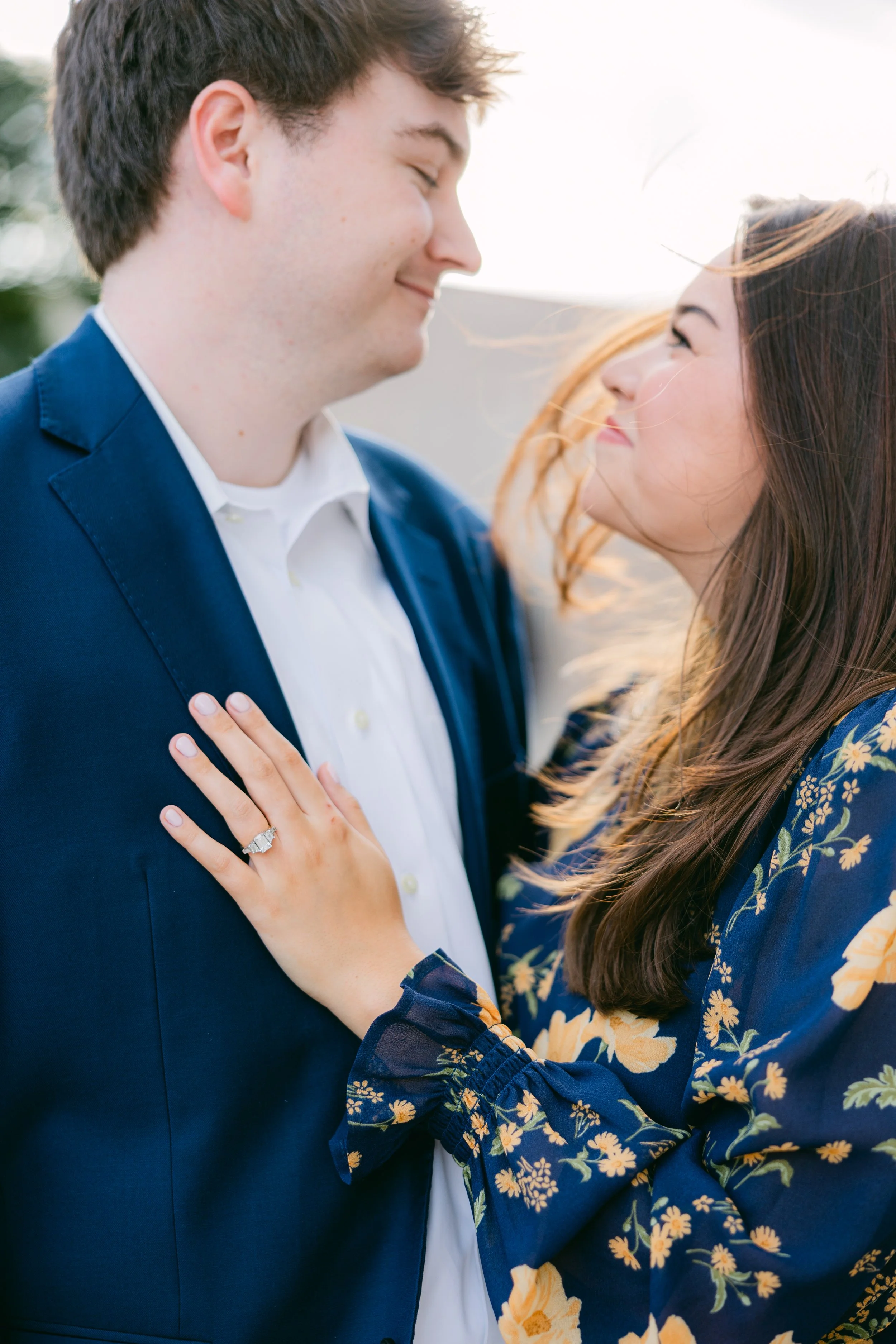 A man and woman face each other closely and smile. The woman has her hand on the man's chest, showcasing her engagement ring. They are outdoors with the sun bright behind them.