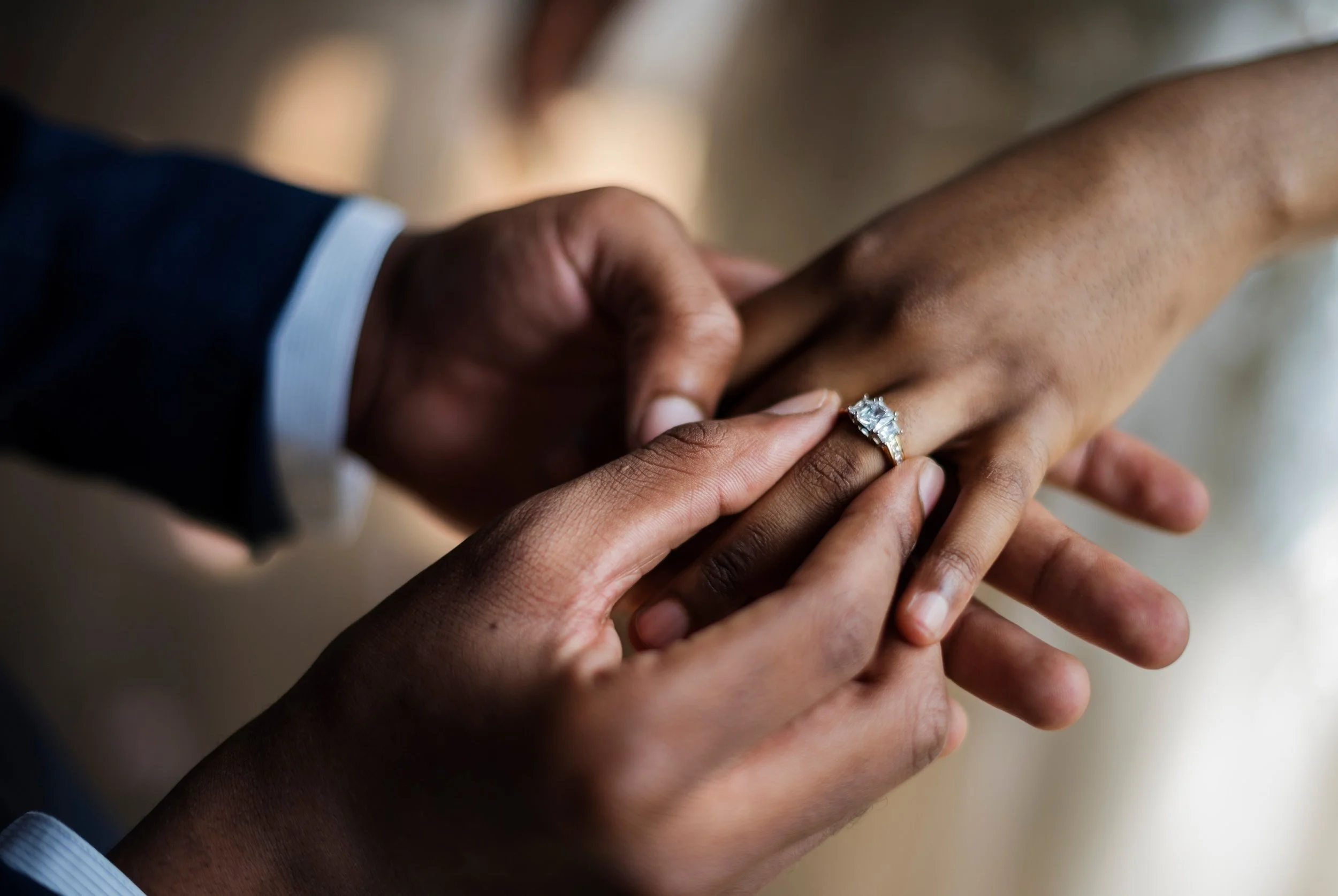 Close-up of a person with dark skin wearing a ring with a large gemstone receiving a ring from another person with lighter skin, in an intimate moment.