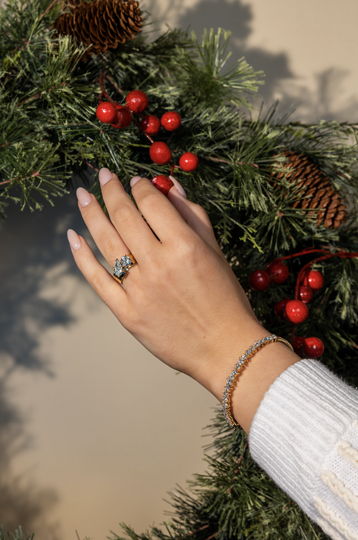 A woman's hand with a diamond ring and a bracelet touching Christmas greenery with red berries and pinecones.