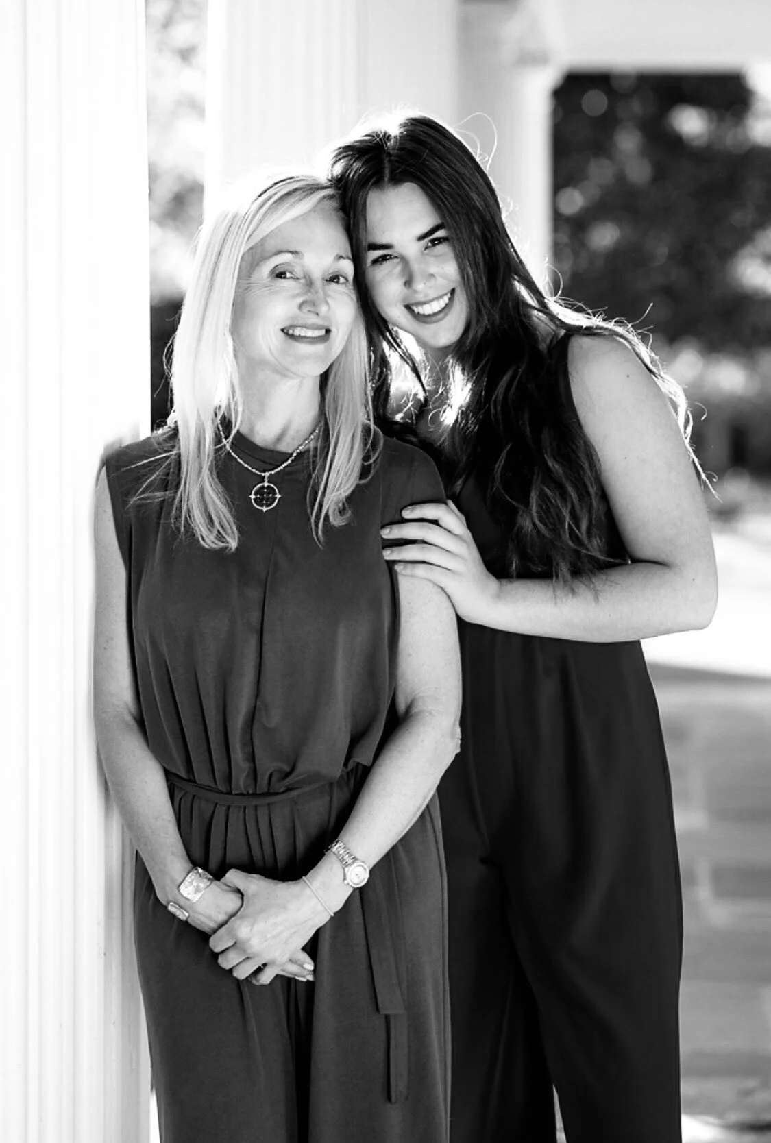 Two women smiling, one older with blonde hair and the other younger with long dark hair, standing outdoors near a pillar with blurred trees in the background.