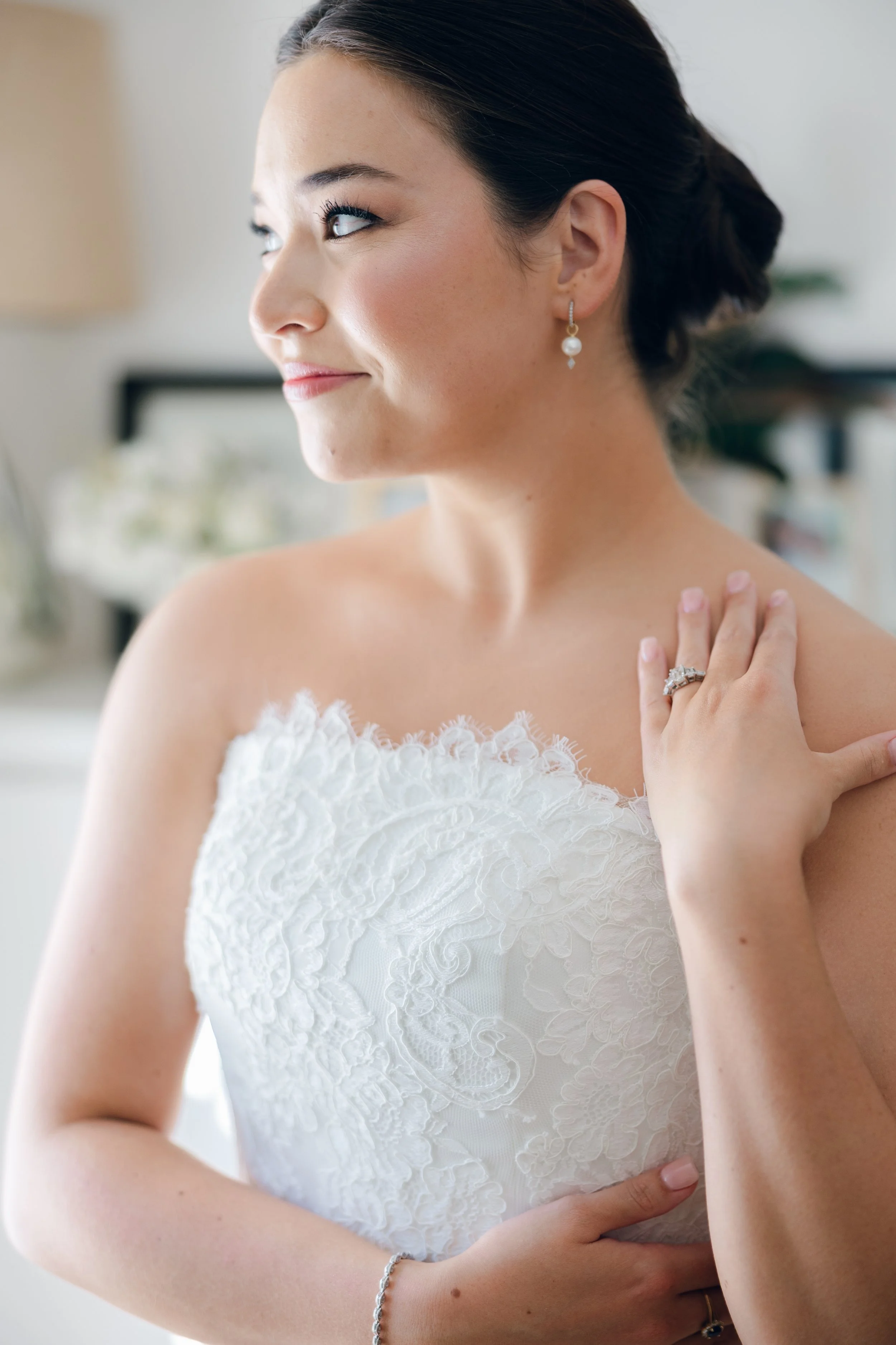 Close-up side profile of a woman in a white dress with lace details, wearing pearl earrings and rings, touching her shoulder with a soft smile and looking away.