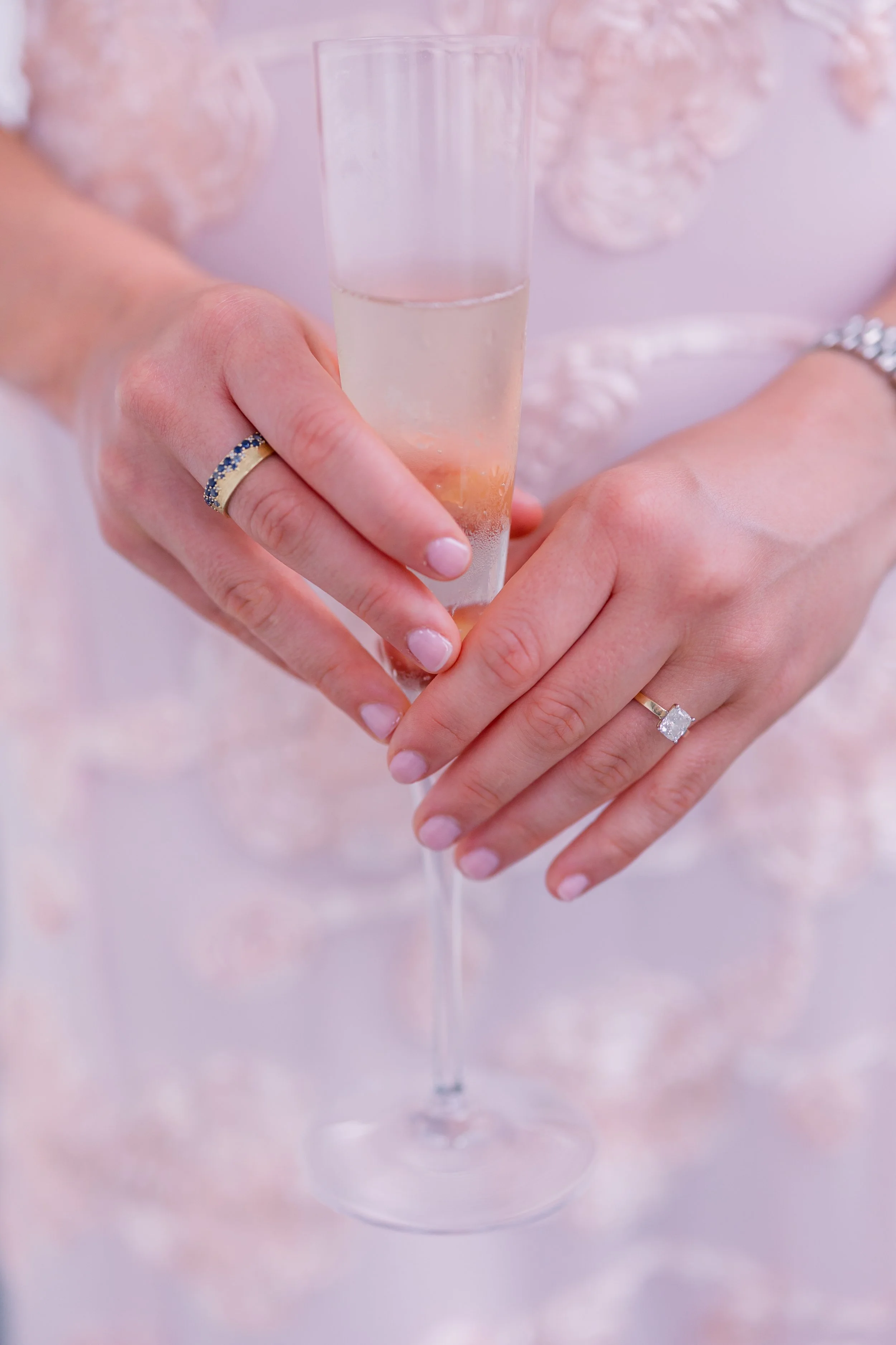 Close-up of a woman's hands holding a champagne flute filled with sparkling wine, with a pinkBackground and floral decorations.