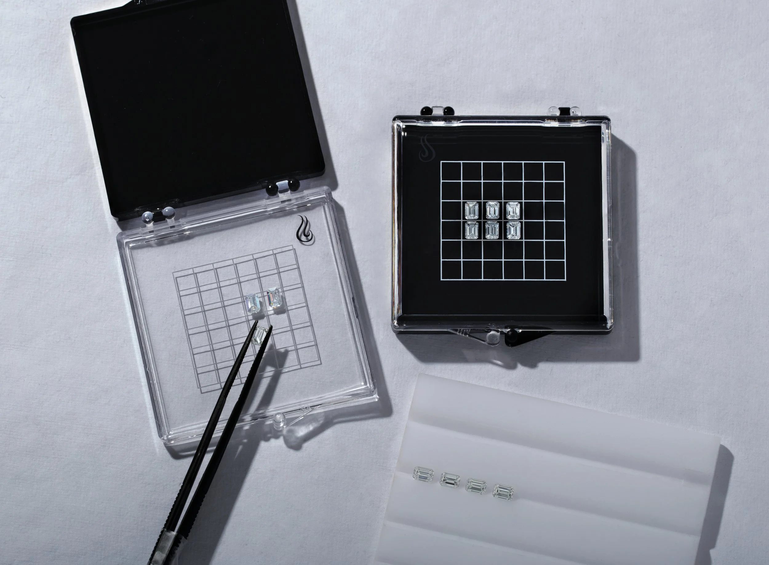 A black and clear acrylic display case with jewelry pieces inside, a pair of tweezers holding a small jewelry piece, and loose jewelry parts on a white surface.