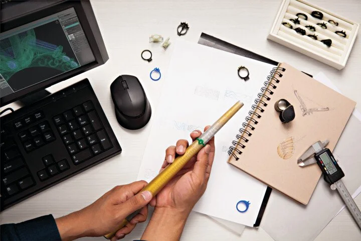 A person working with jewelry design tools on a white desk, including a sketchpad with jewelry sketches, a digital caliper, rings, and a computer with 3D modeling software.