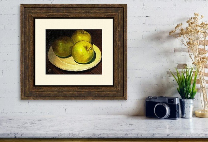 A framed still life painting of three green apples on a white plate hung on a white brick wall, with a potted plant, camera, and dried flowers on a shelf below.