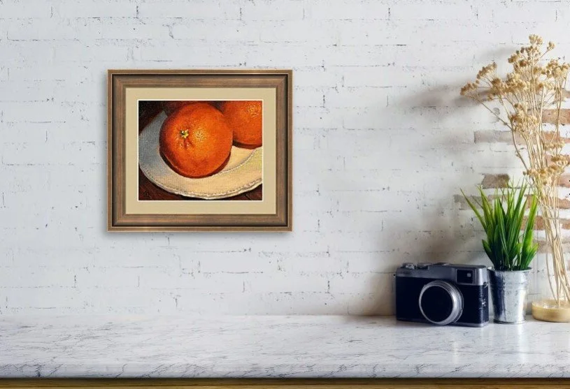 A framed still life painting of three oranges on a white plate hangs on a white brick wall, with a camera, potted green plant, and dried flowers on a white shelf below.
