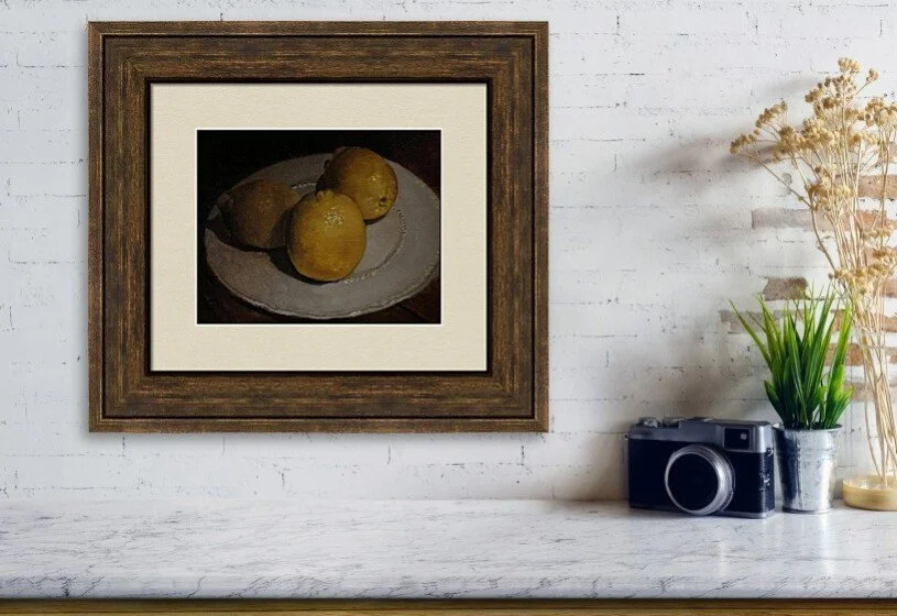 A framed painting of three yellow lemons on a white plate hangs on a white brick wall above a wooden shelf, which holds a vintage camera, a potted plant, and dried flowers.