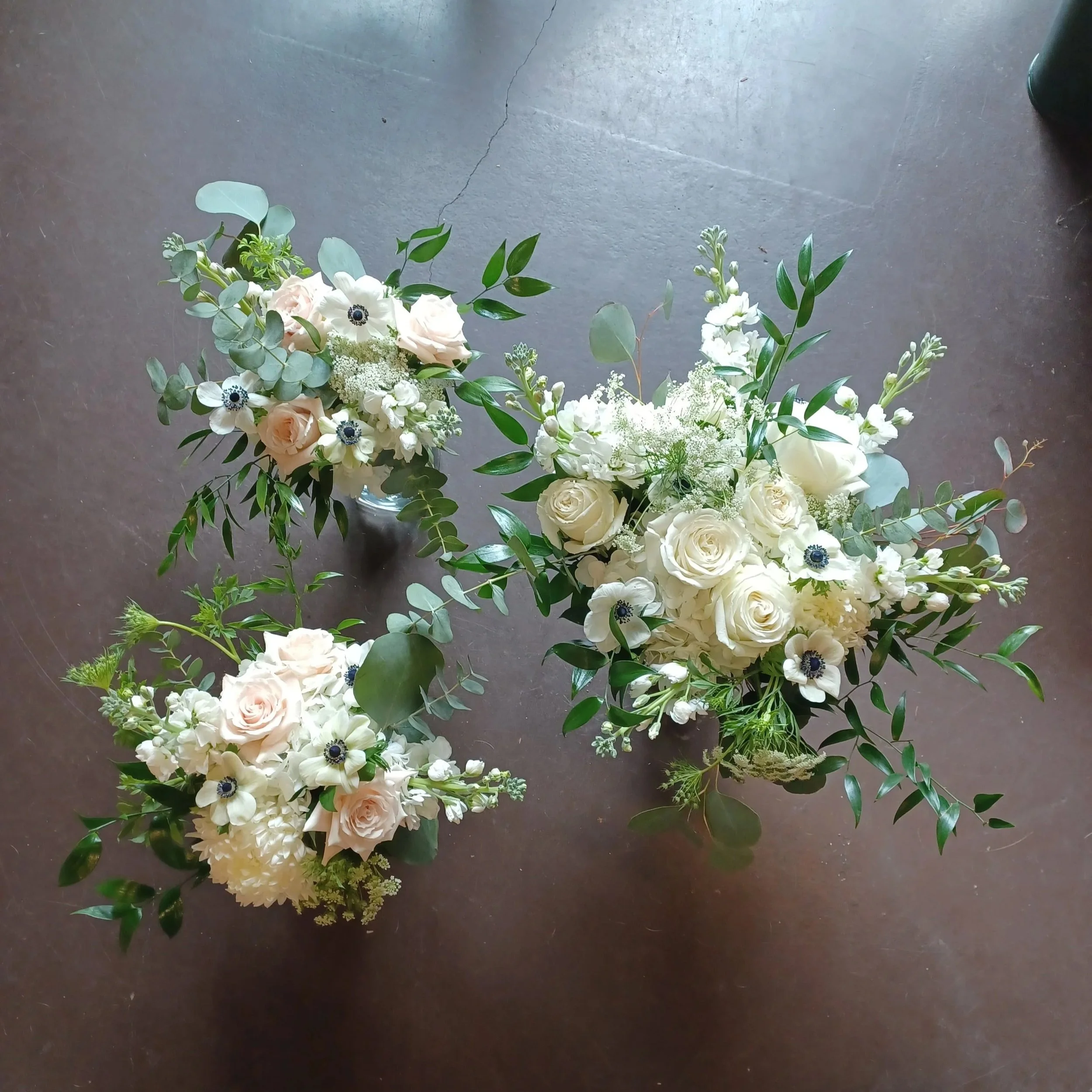 Three arrangements of white flowers with greenery on a dark surface, viewed from above.