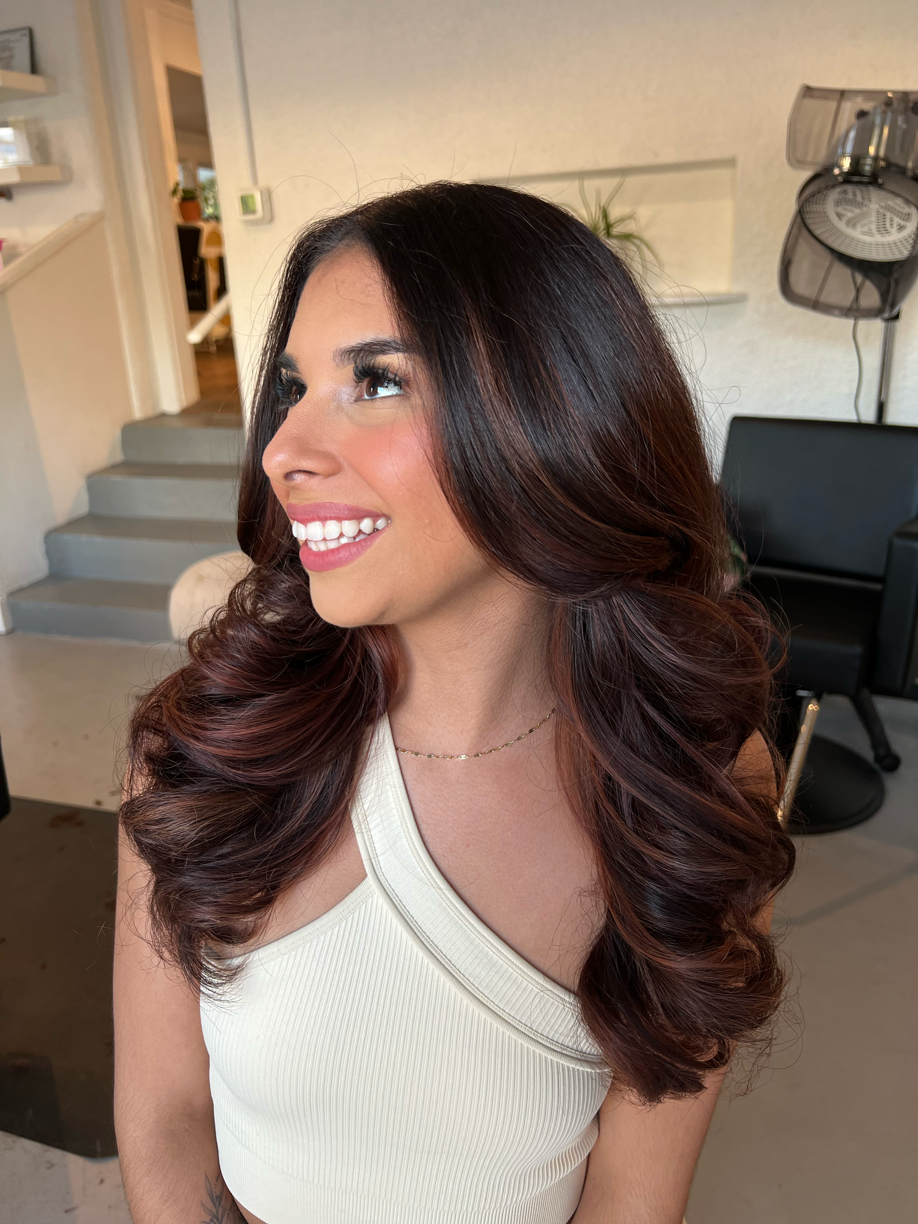 Woman with long, wavy brown hair, wearing a white tank top and a delicate gold necklace, smiling and looking to her left inside a modern home.