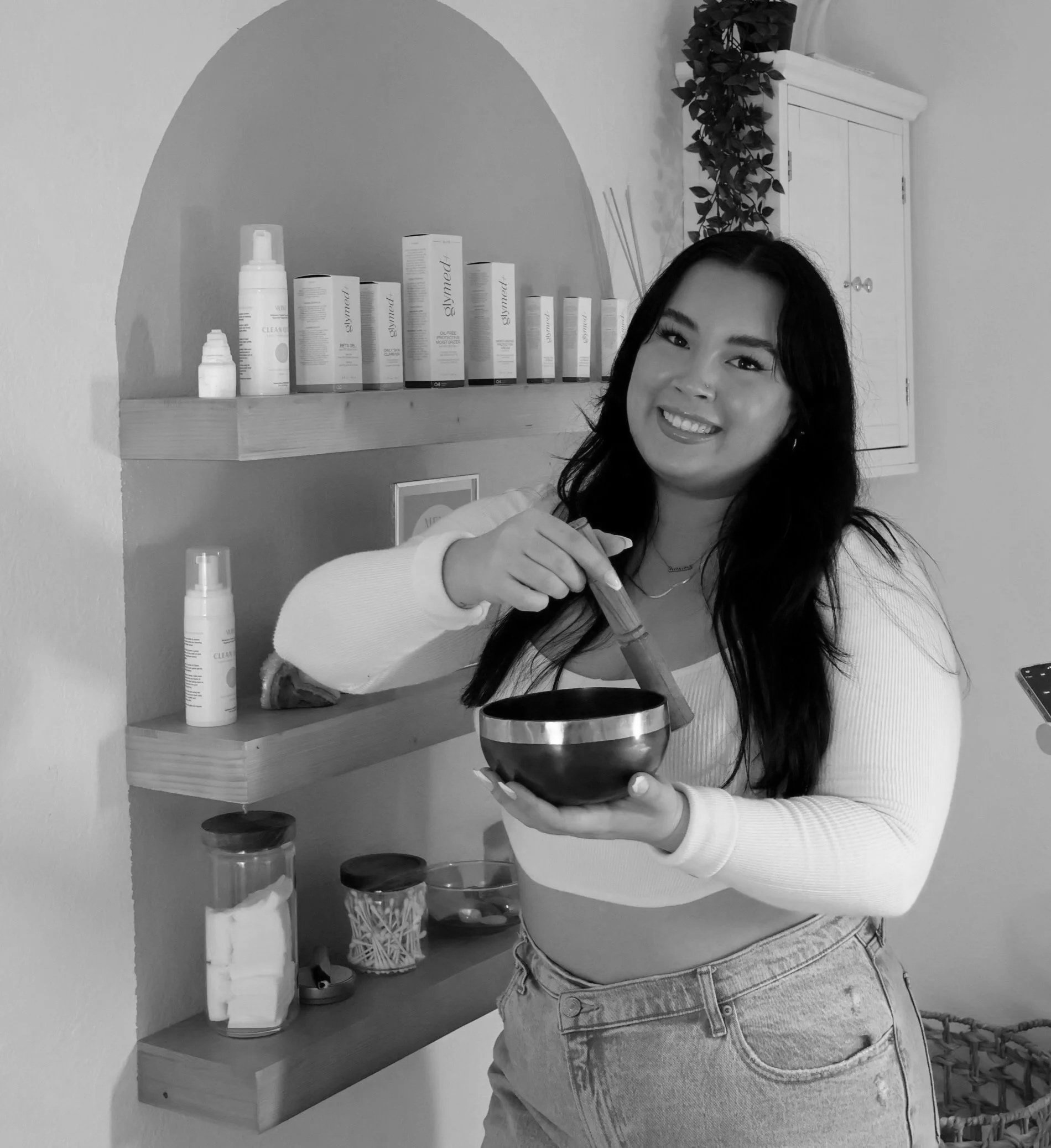 A woman with long black hair smiling and holding a singing bowl while using a stick to play it in a cozy room with shelves of skincare products and decorative items.