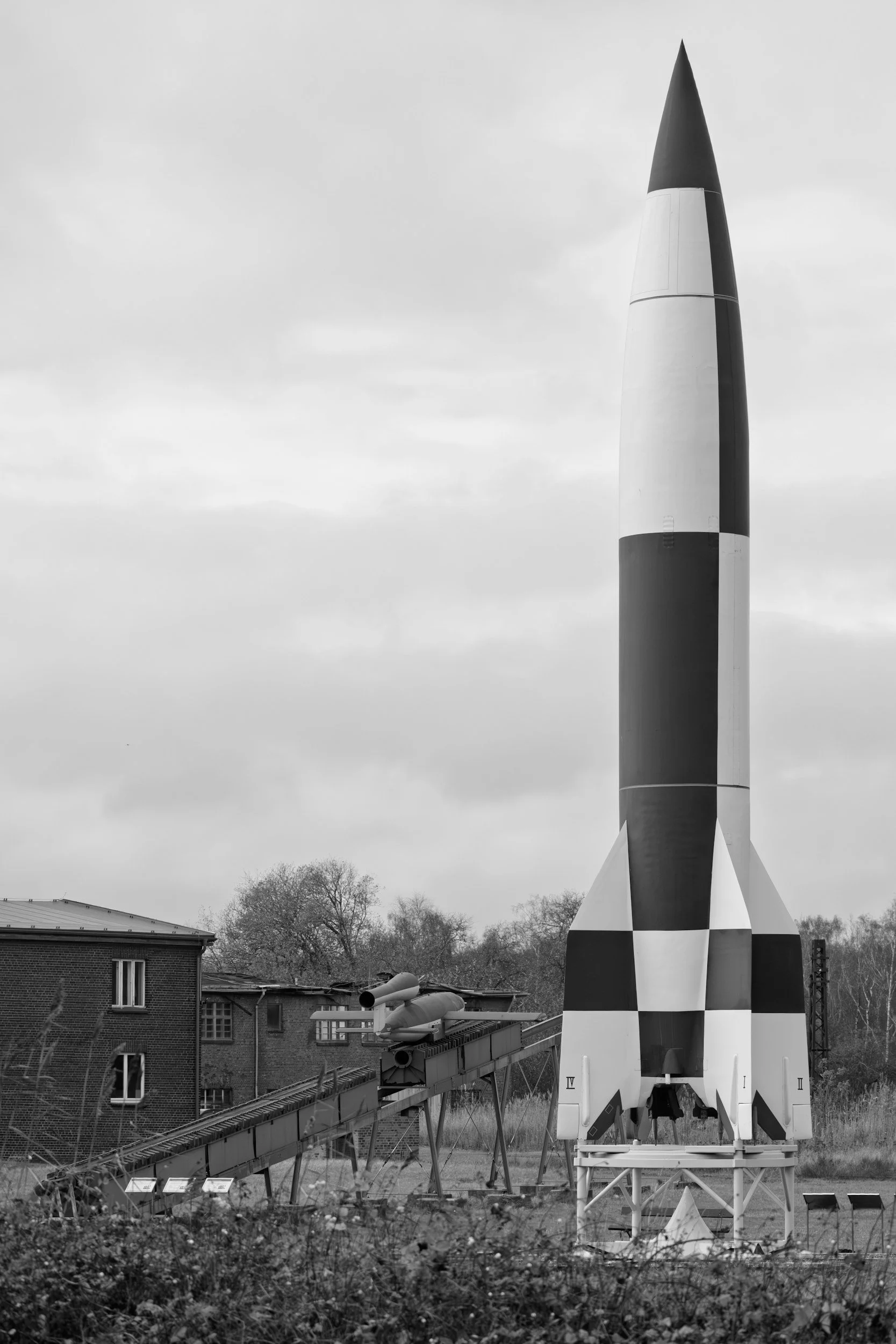 A black and white photo of a model rocket standing upright outdoors with a small building and leafless trees in the background.