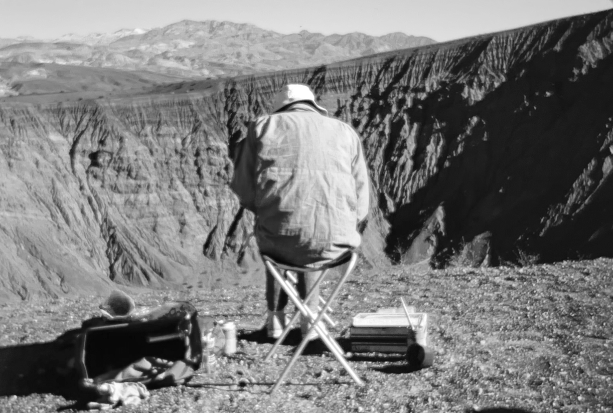 A person wearing a hat sits on a foldable stool near the edge of a canyon, with a suitcase and other items on the ground, overlooking a vast landscape of rugged mountains and valleys.
