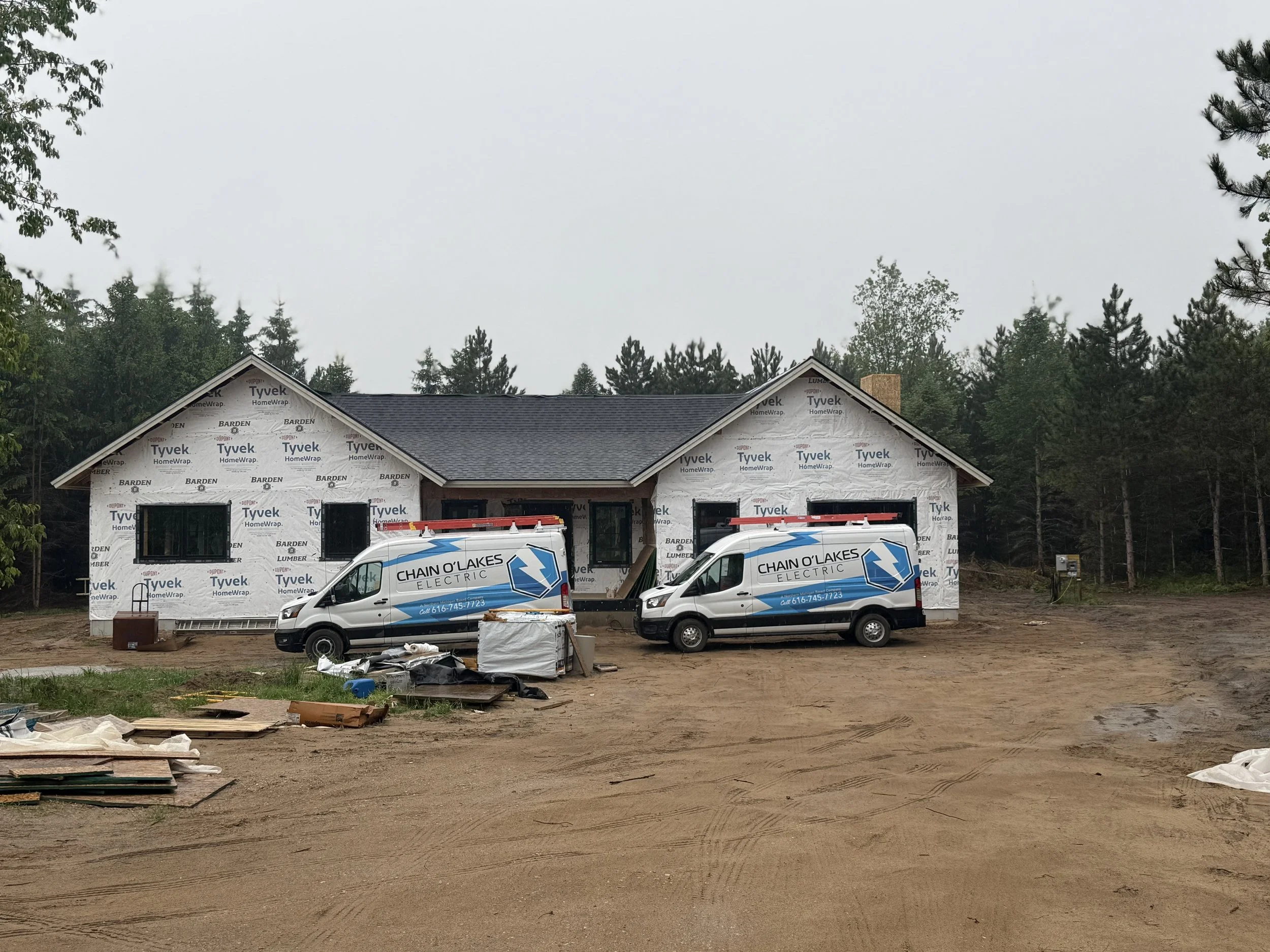 A house under construction with two Chain O'Lakes Electric service vans parked in front. The house is wrapped with Tyvek HomeWrap and surrounded by trees.
