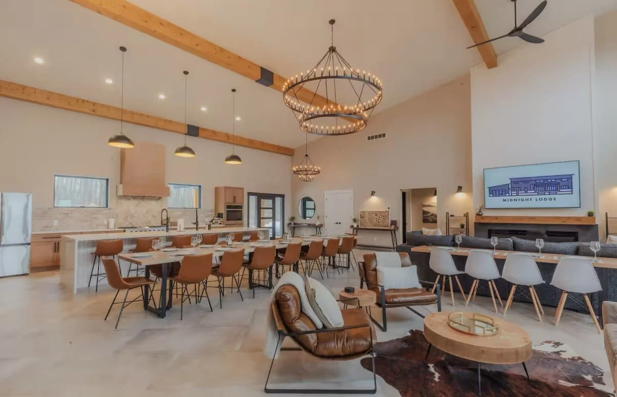 Open-concept kitchen and living room in a modern lodge with wood beams, pendant lighting, a chandelier, bar seating, a large dining table, a leather armchair with pillows, a sectional sofa, a large flat-screen TV, and decorative elements.