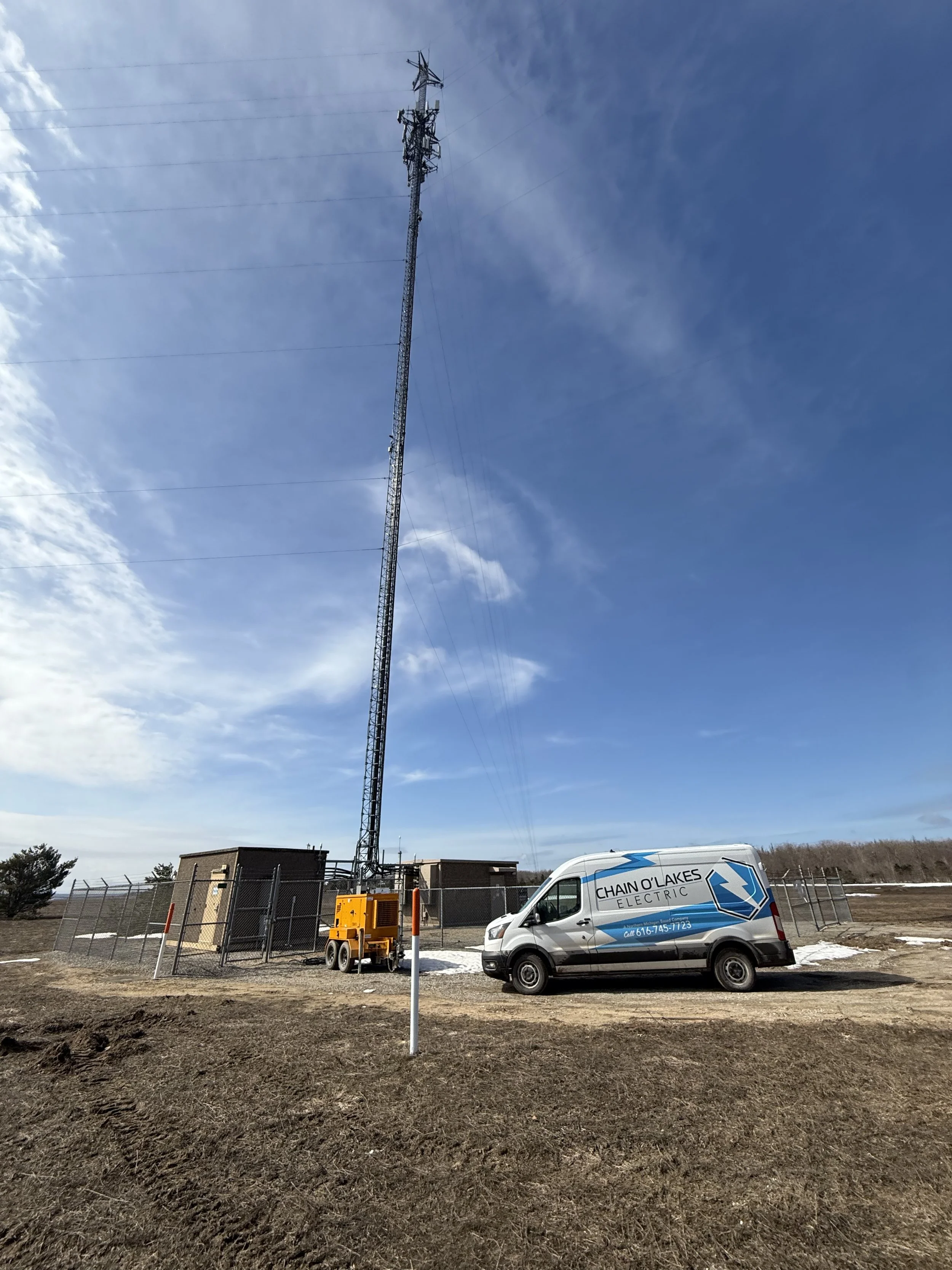 A tall radio tower with multiple antennas and wires, located near a fenced area with a utility building, a yellow generator, and a white electric service van labeled 'Chain O Lakes Electric', under a partly cloudy blue sky.