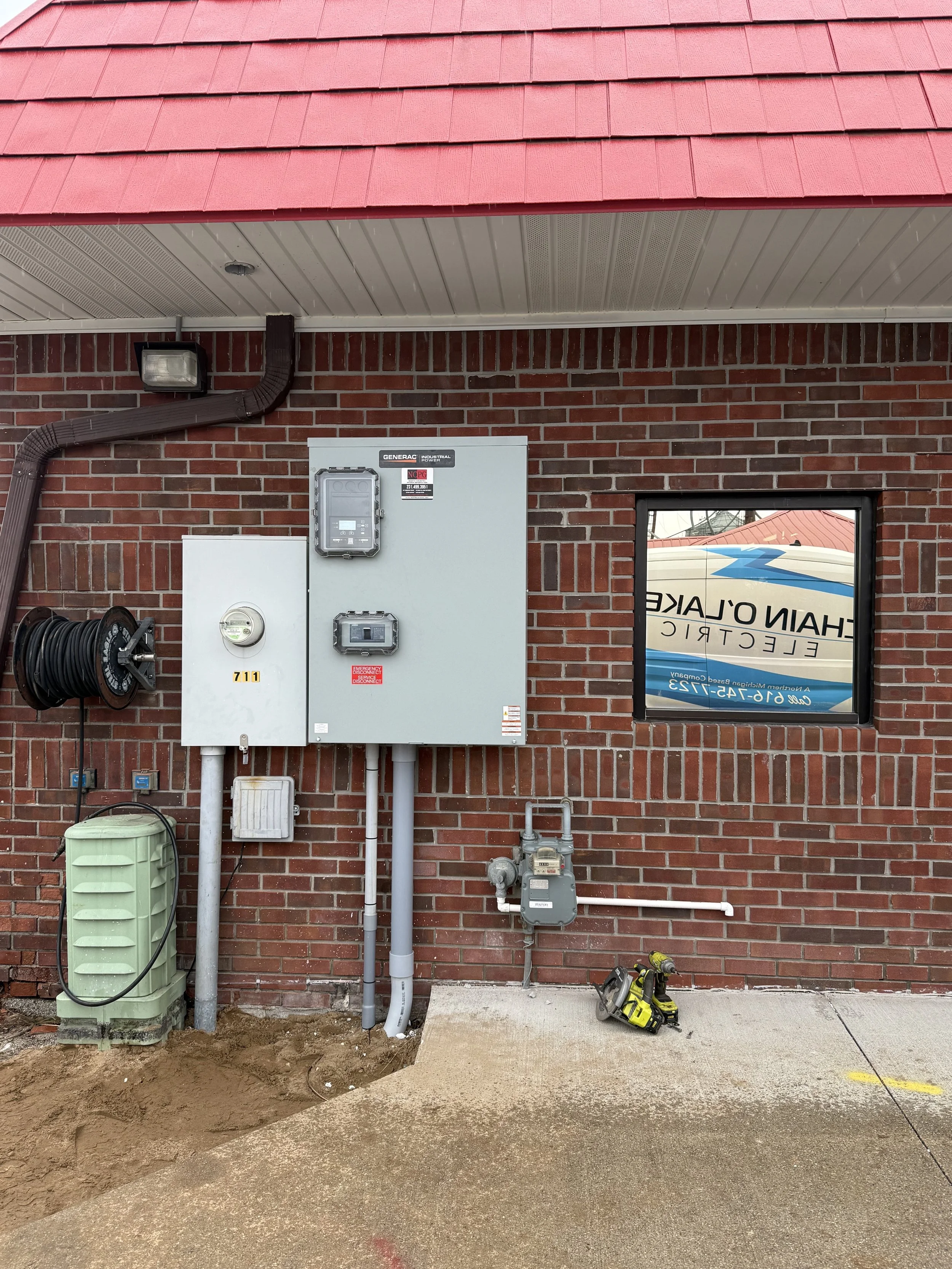 Exterior view of a brick building with utility and electrical boxes, a hose reel, a green water tank, a window with a reflection of a boat, and a cordless drill on the ground.