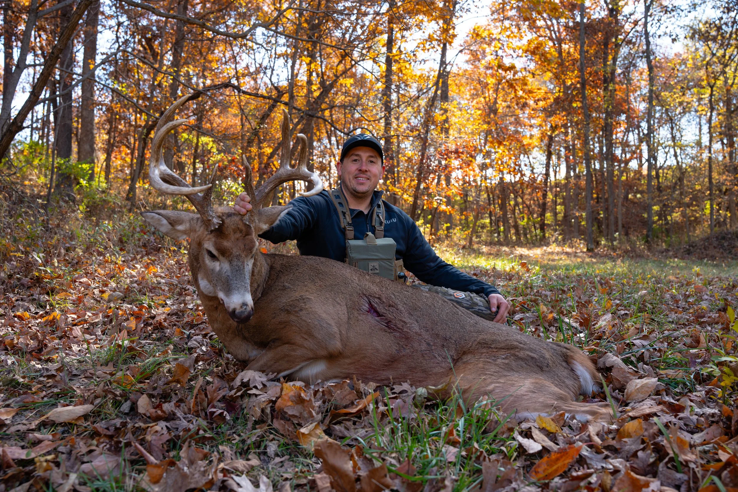 A man kneeling in a forest with autumn foliage, holding the antlers of a large deer he has hunted, which is lying on the ground.