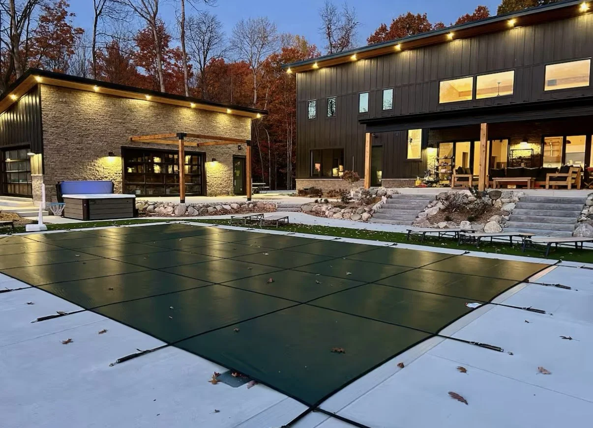 Backyard with outdoor furniture, a green covered hot tub, and an open-air basketball hoop, surrounded by illuminated modern houses and fall foliage.