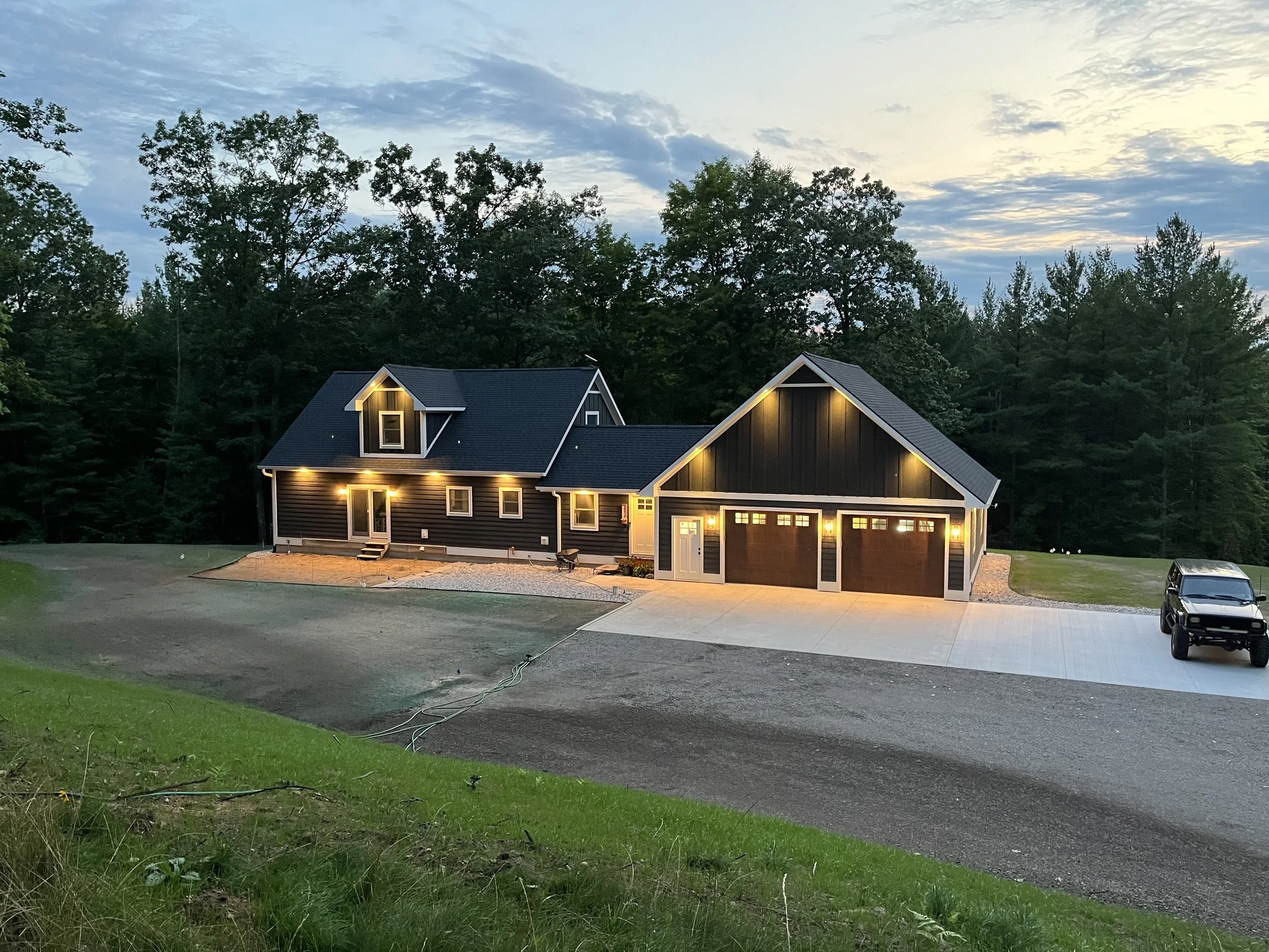 A newly constructed black house with a garage illuminated by exterior lighting at dusk, surrounded by trees and a spacious driveway with a parked black vehicle.