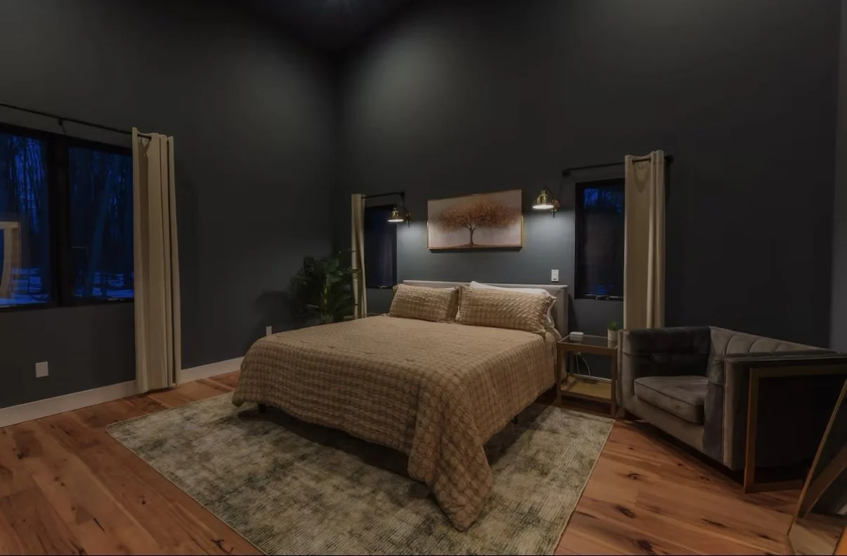 A bedroom with dark gray walls, a large bed with a tan checkered quilt, two matching pillows, and wall-mounted lamps. There are two windows with beige curtains, a framed tree artwork above the bed, a nightstand with a plant and clock, a gray armchair