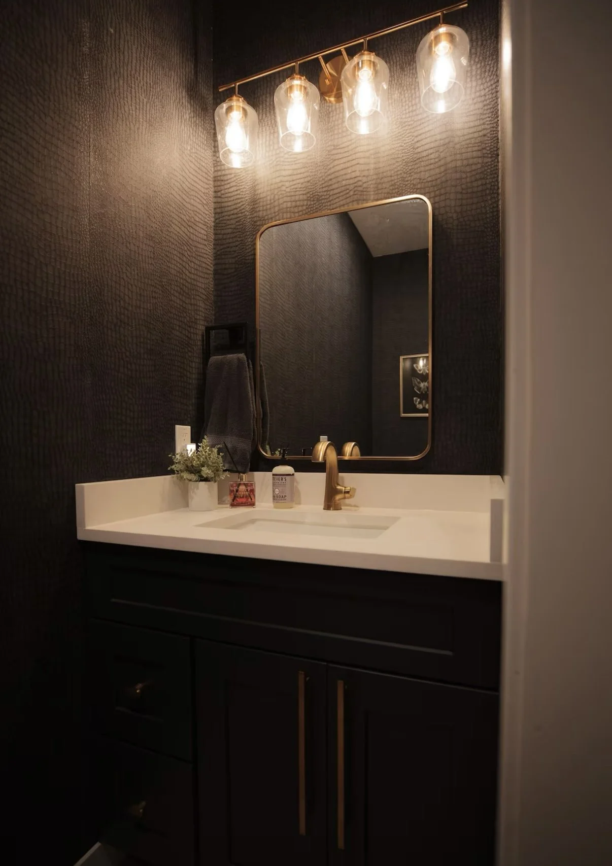 A bathroom with dark textured walls, a white countertop, and a black vanity with gold handles. There is a gold faucet, a mirror, and four hanging glass light fixtures above the sink. Items on the counter include a small plant, soap, and other toiletr