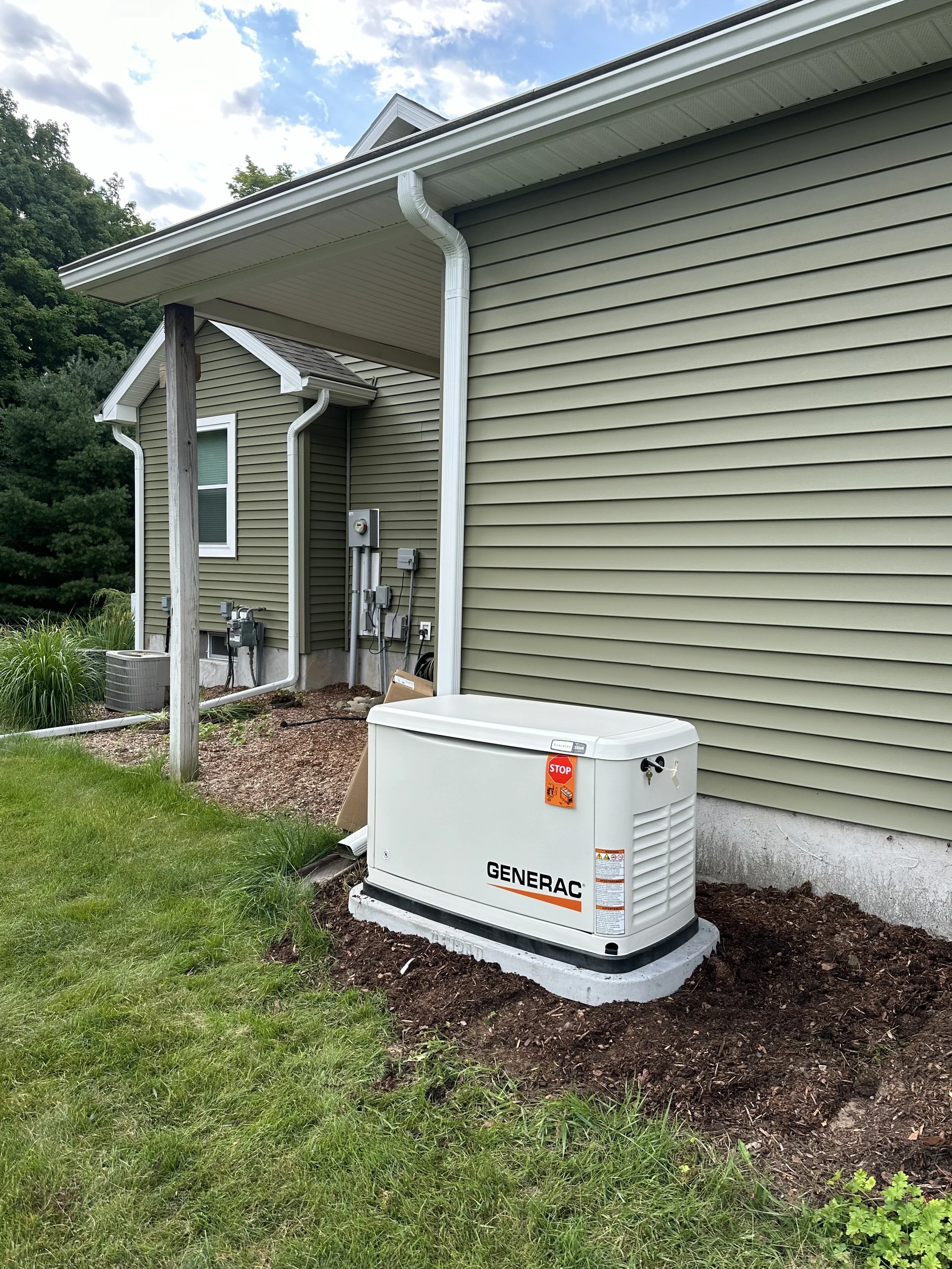 A white Generac generator installed outside a house with green siding, on a concrete pad, near a grassy lawn and under a small covered porch.