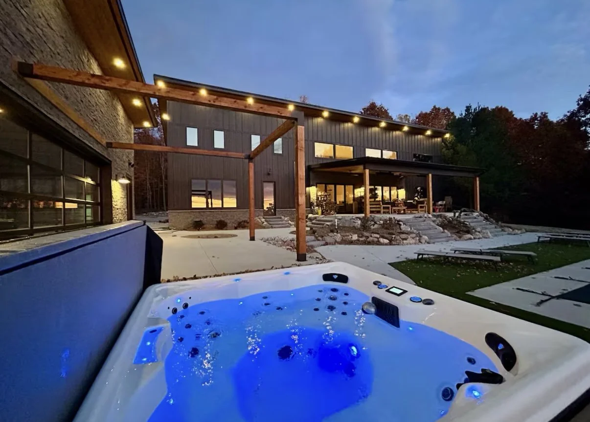 Image of a house at dusk featuring a hot tub with blue lights in the foreground, a spacious patio, wooden pergola, large windows, and trees with fall foliage in the background.