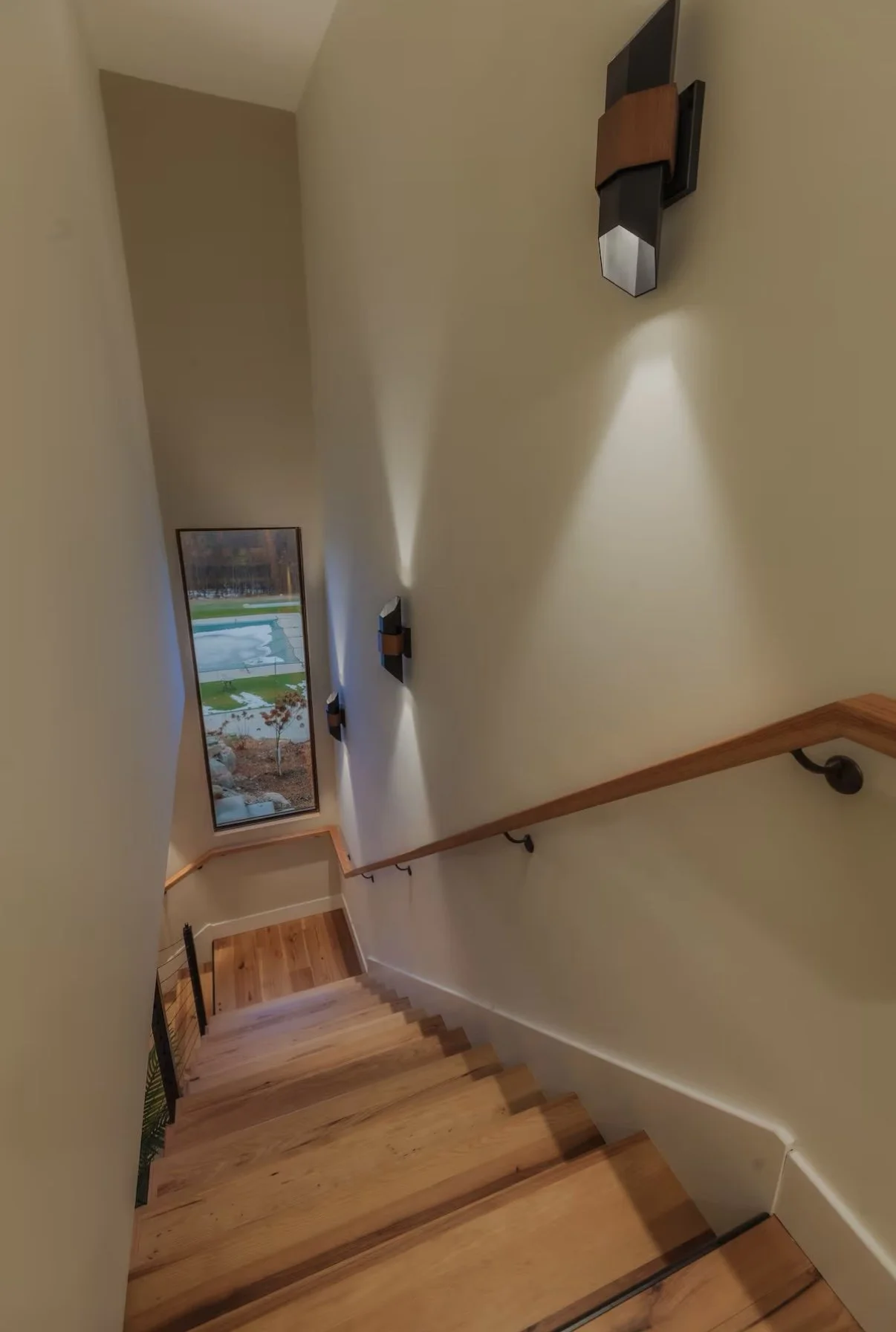 View of a staircase descending to a landing with a tall window showing an outdoor yard and pool. Wall-mounted lights cast light on a beige wall, and there's a wooden handrail along the staircase.