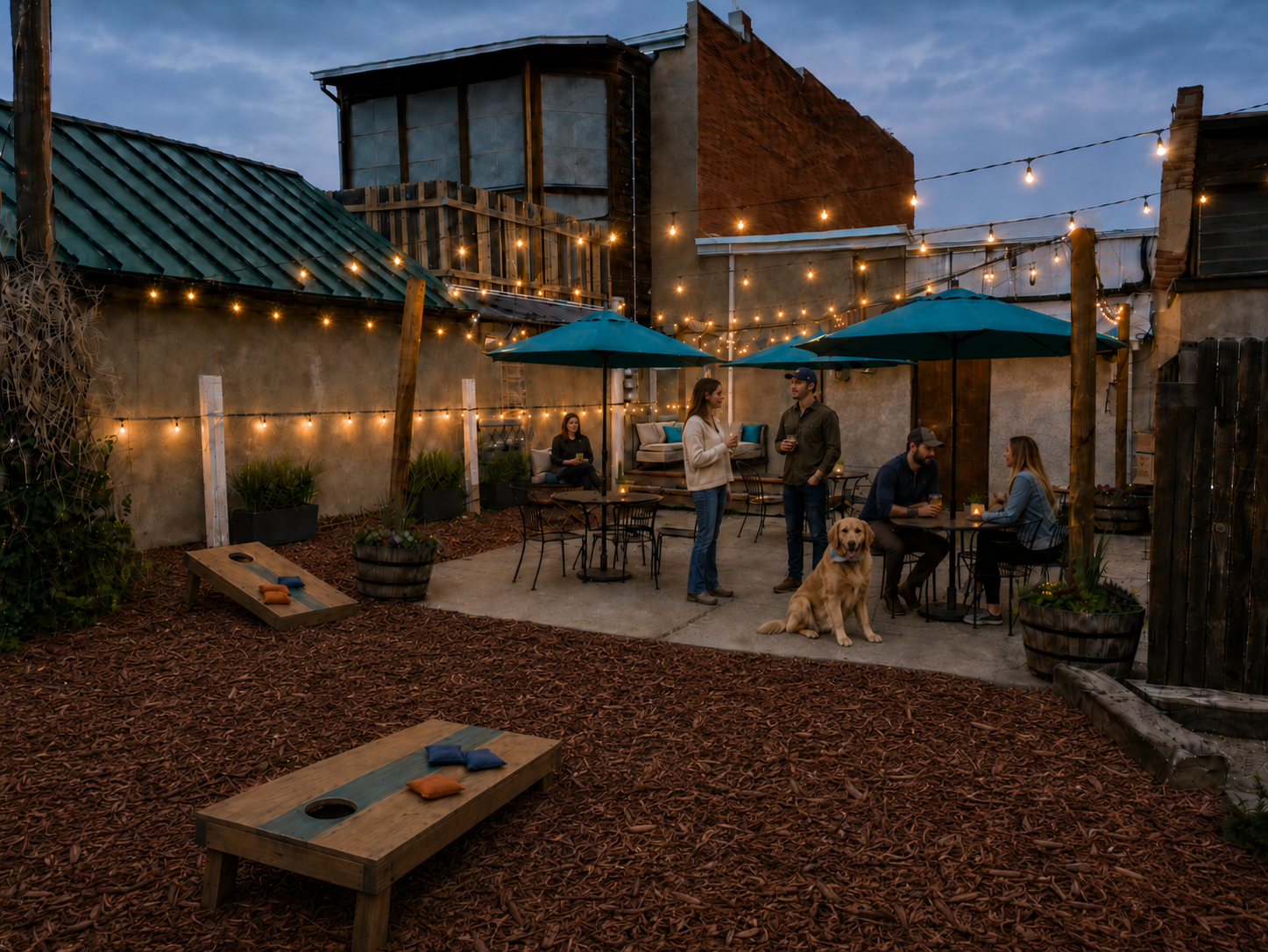 An outdoor patio with ambiance - string lights, people socializing, and a dog friendly space. There are tables with teal umbrellas, cozy seating, and a cornhole game on mulch grounds.